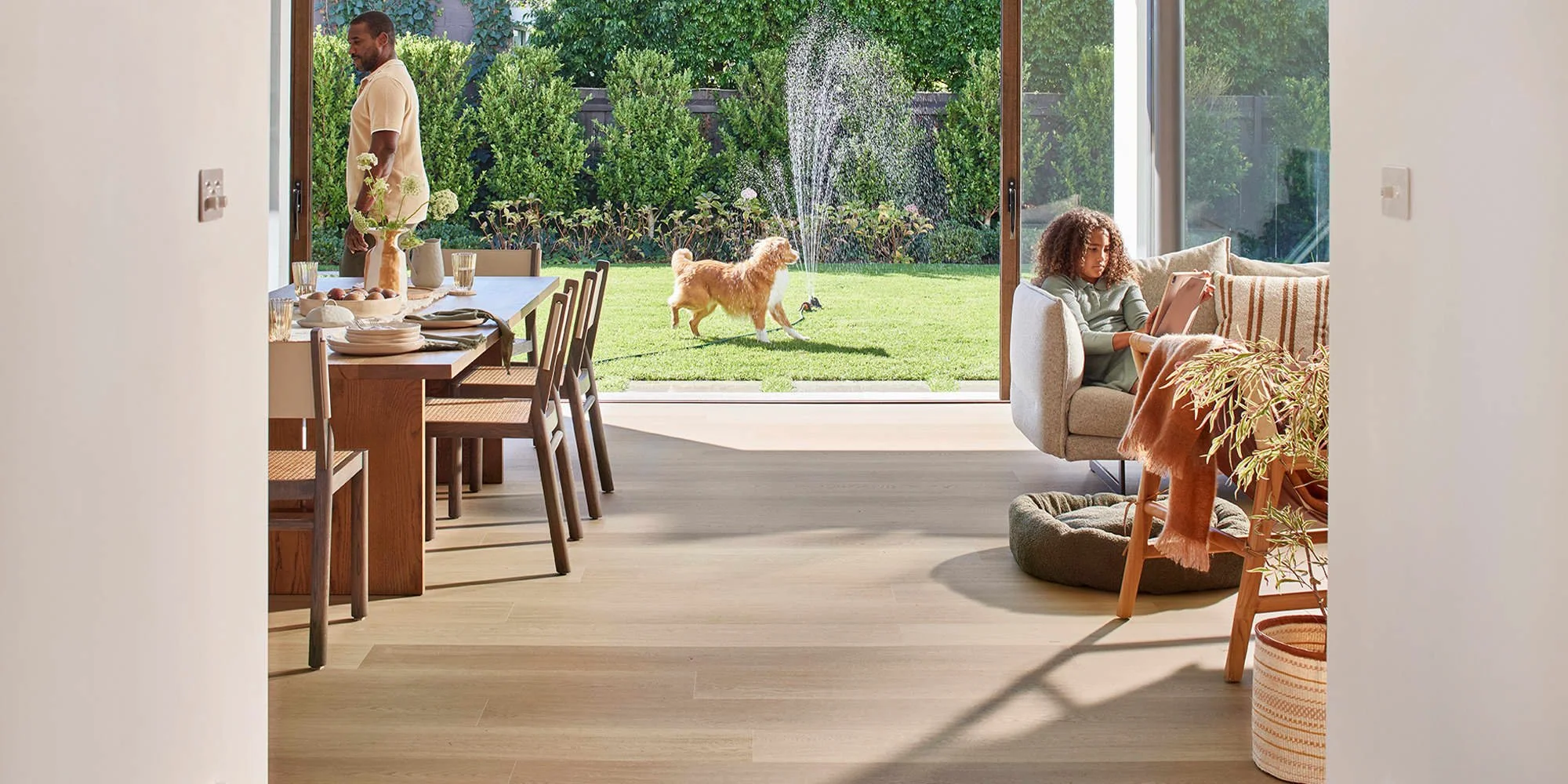 Living room with a view of a backyard, a woman sitting on a sofa using a tablet, a man standing near the dining table, a dog playing with a toy fountain, and decor including a potted plant and dining set.