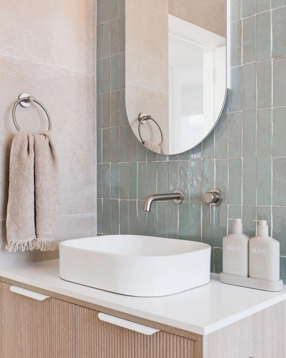 Bathroom with beige and green tiled wall, oval mirror, beige towel on towel ring, modern faucet, white vessel sink, and two white soap bottles on a tray.