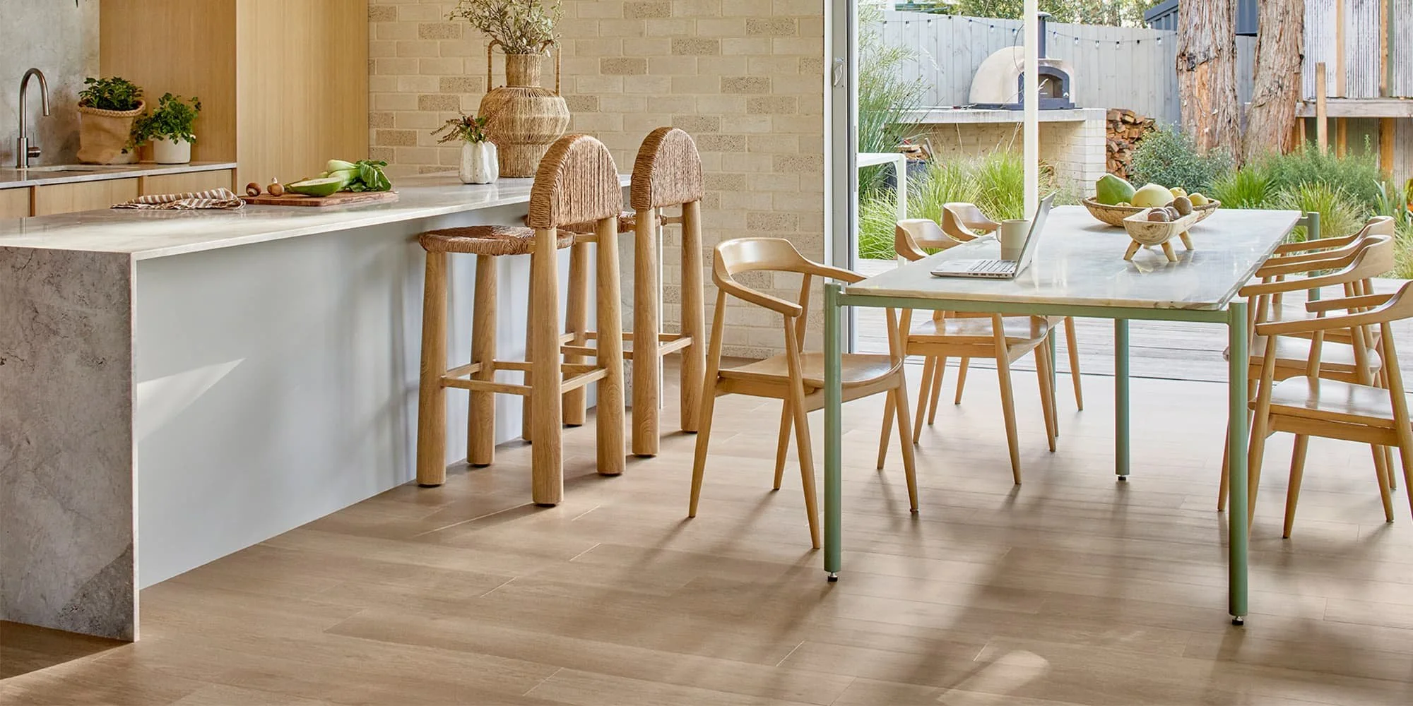 Modern kitchen and dining area with a white marble island, wooden barstools, a glass dining table with wooden chairs, and a view of a backyard with a pizza oven and outdoor seating.
