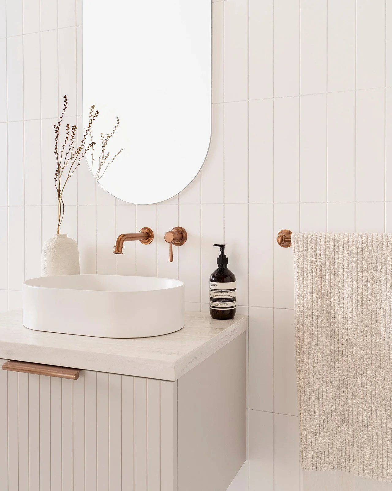 Minimalist bathroom with white tiles, oval ceramic sink, brass wall-mounted faucet, black soap dispenser, beige towel on rod, and a vase with dried branches in the corner.