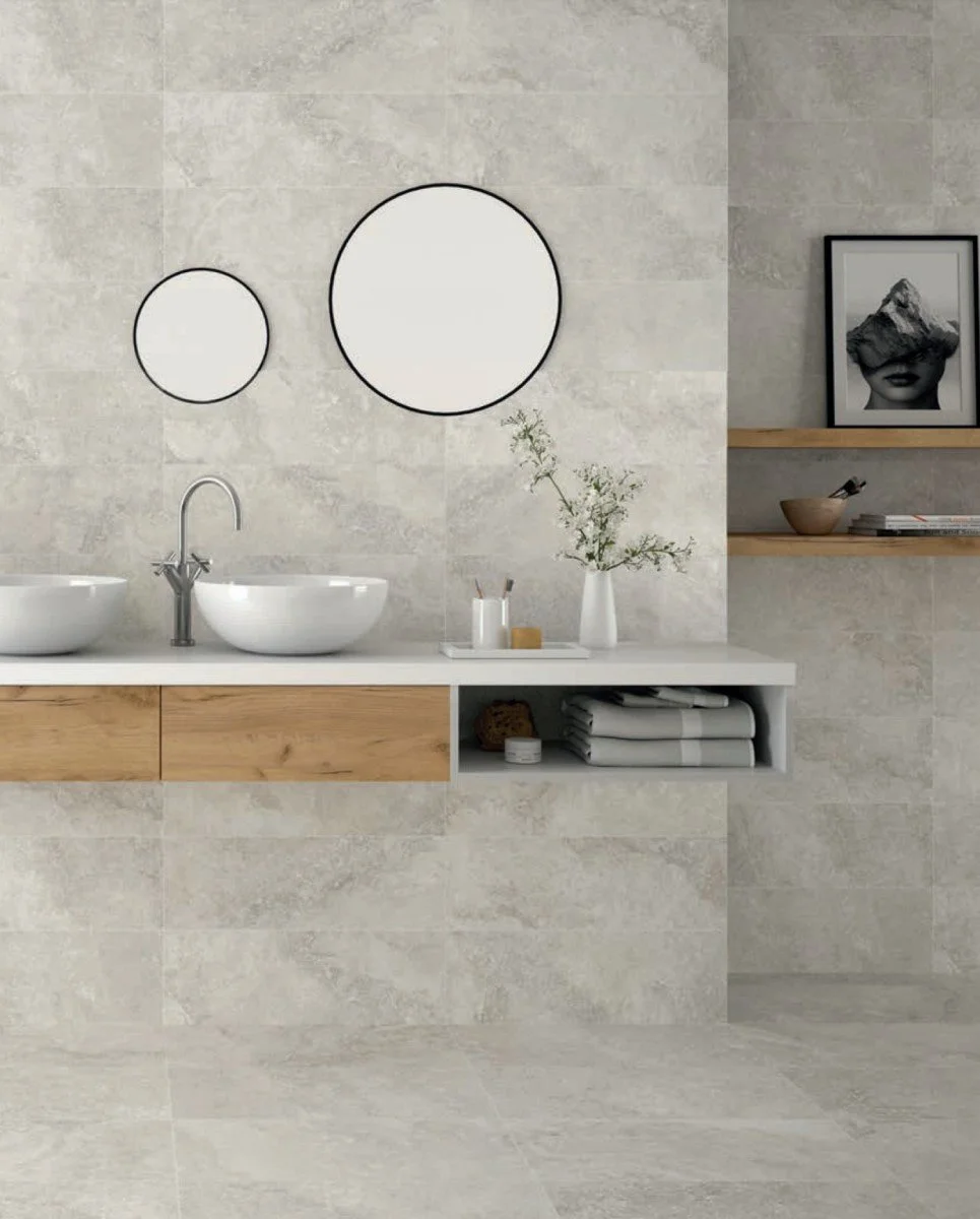 Modern bathroom with a white floating vanity, two white vessel sinks, and a chrome faucet. Above the sinks are two round mirrors. To the right, a framed black-and-white art piece and two wooden shelves hold small decor items and rolled towels. A vase