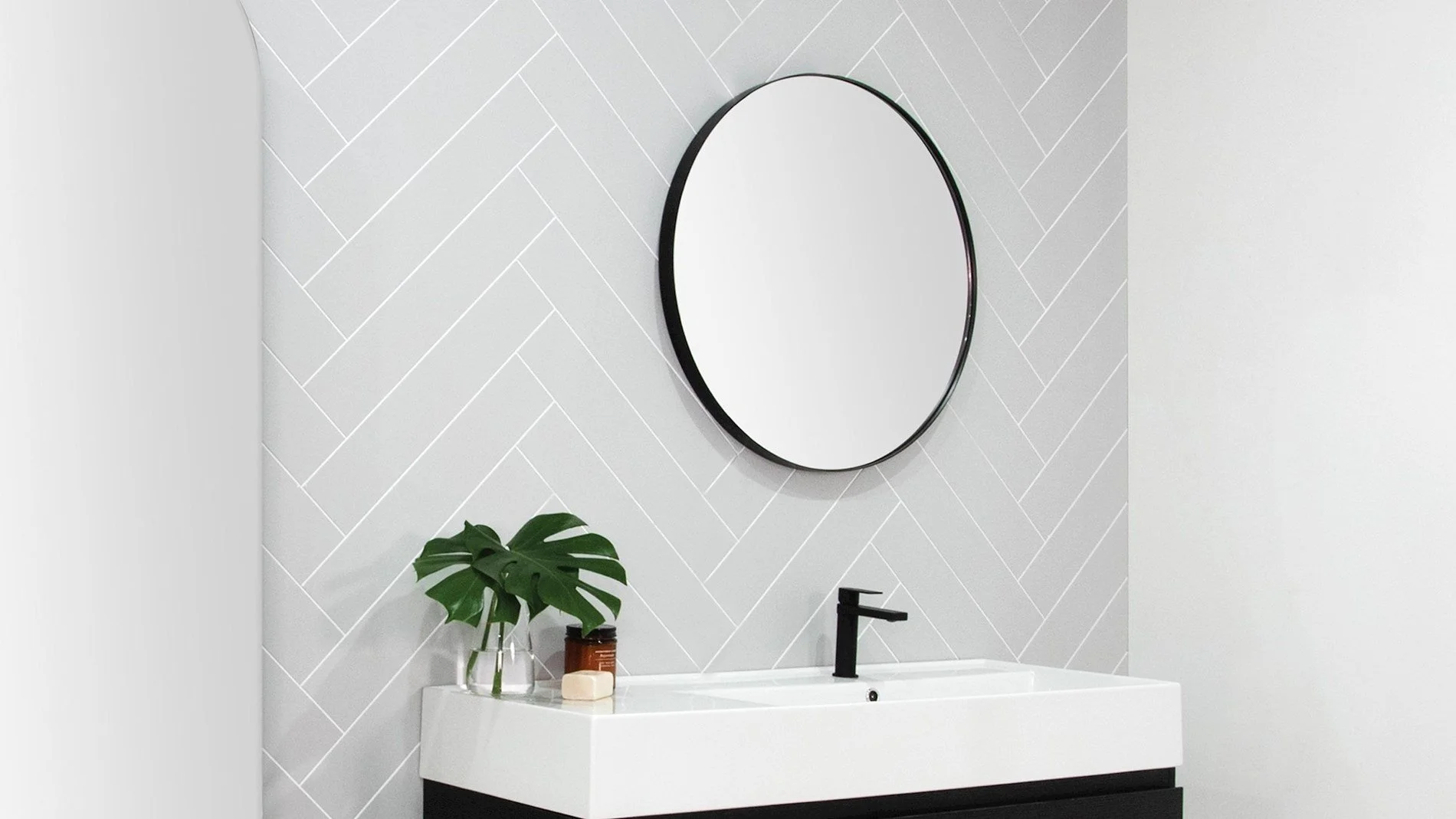 Modern bathroom vanity with a white sink, black faucet, large round mirror, and green plant in a glass vase against a white herringbone tile wall.