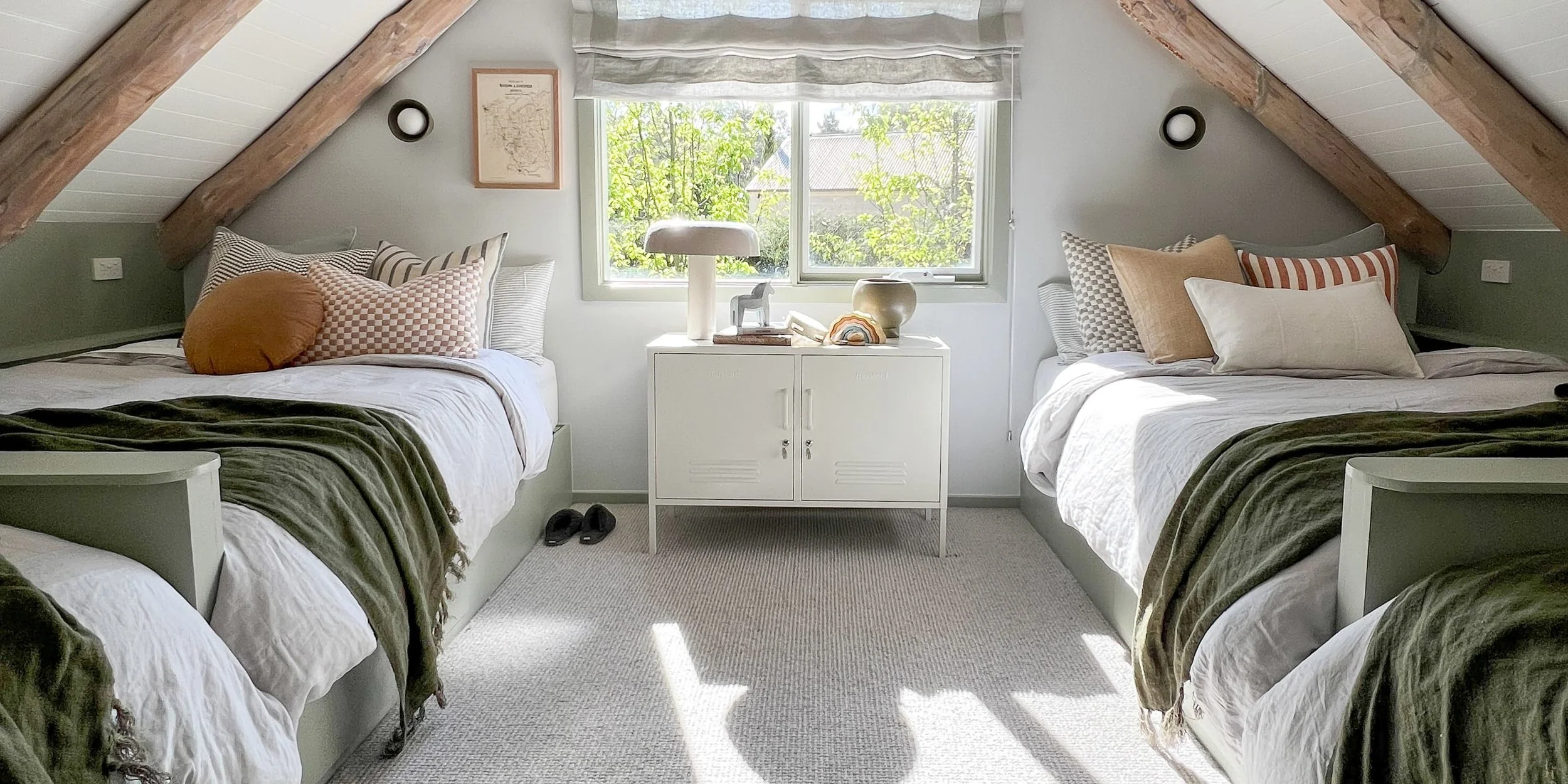 An attic bedroom with two beds, decorated with pillows and blankets, a white cabinet between the beds, a window with greenery outside, and wooden beams on the ceiling.