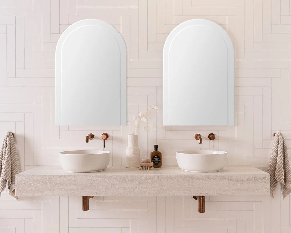 Bathroom with dual white vessel sinks, arched mirrors, beige countertop, and two towels hanging on either side.