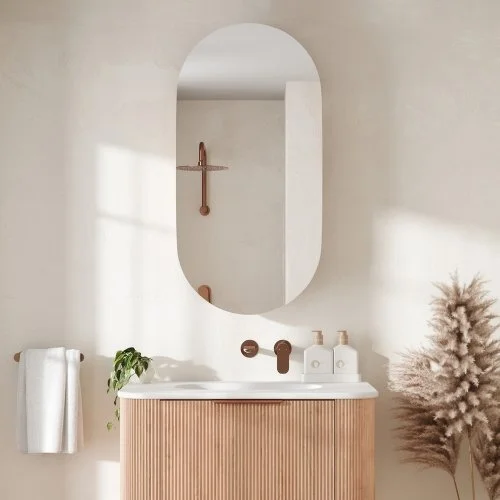 Modern bathroom with oval mirror, wooden vanity, white sink, and minimal decor including a towel, plant, and dried flowers.