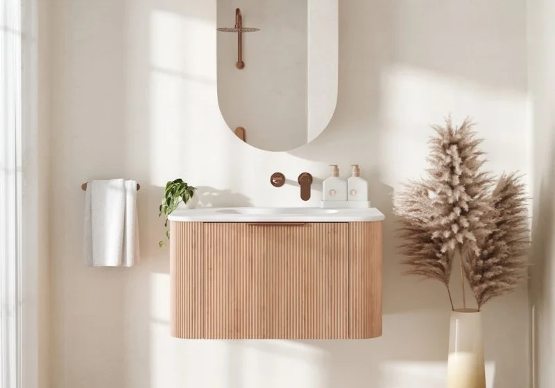 Modern bathroom with a wooden vanity, oval mirror, wall-mounted faucet, potted plant, towel on a wall hook, and pampas grass in a white vase.