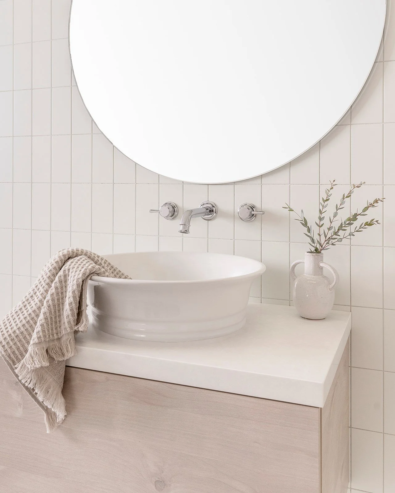 A white ceramic vessel sink on a light wood vanity with a beige towel hanging on the side, a small white vase with green leafy branches, tiling, and a large round mirror above.