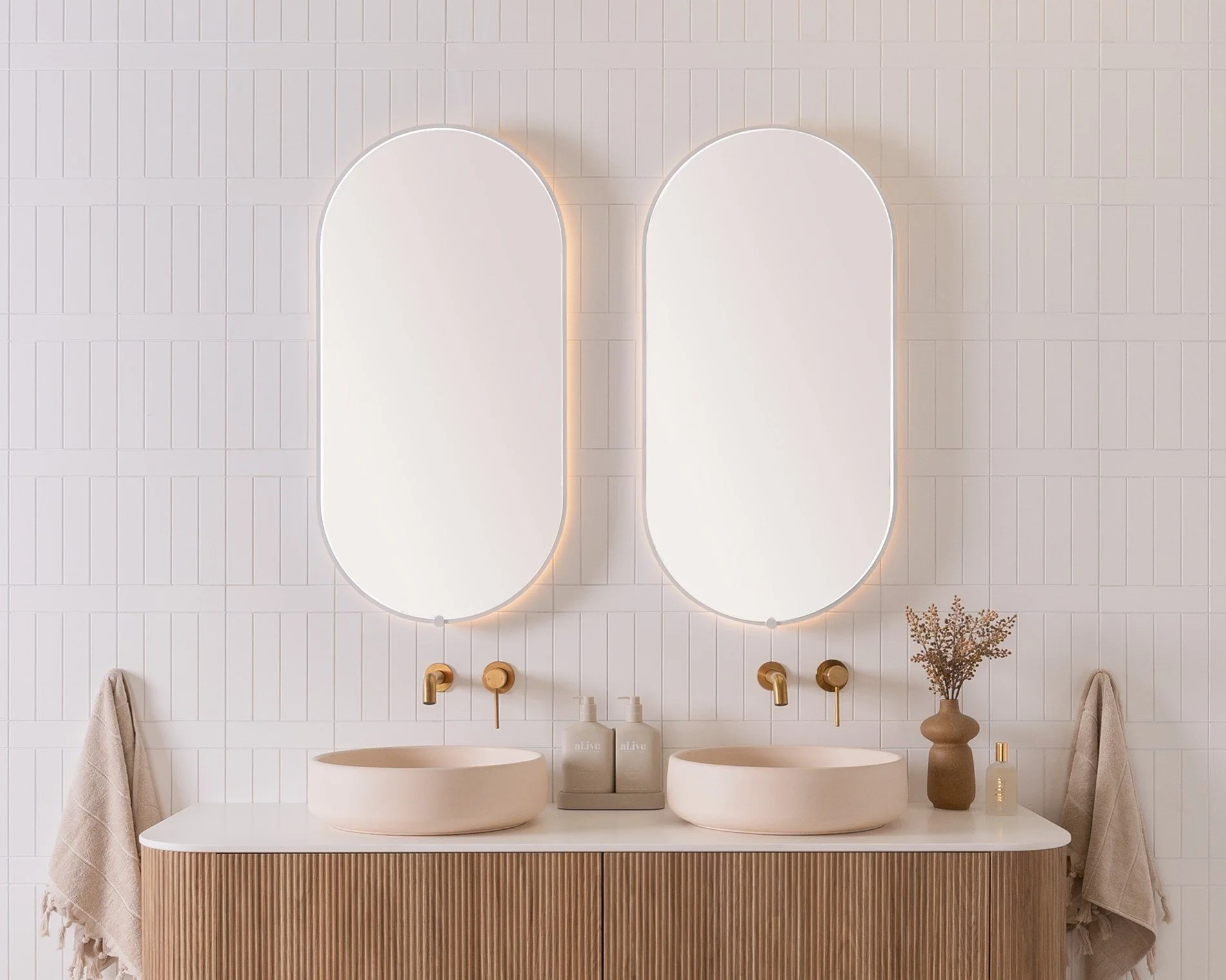 Modern bathroom vanity with two oval above-counter sinks, large vertical rectangular mirrors with rounded tops, beige towels on either side, and decorative items including a vase with dried flowers, soap dispensers, and a small perfume bottle.