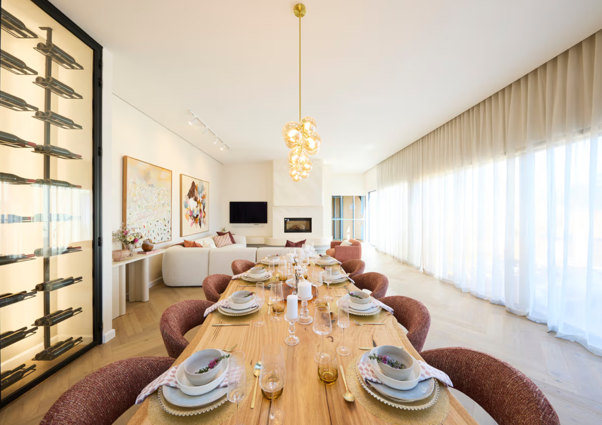 Bright, modern dining room with a long wooden table set for a meal, with wine glasses, candles, and place settings. Large windows with sheer curtains provide natural light, with a white sofa and wall art in the background. A contemporary chandelier h