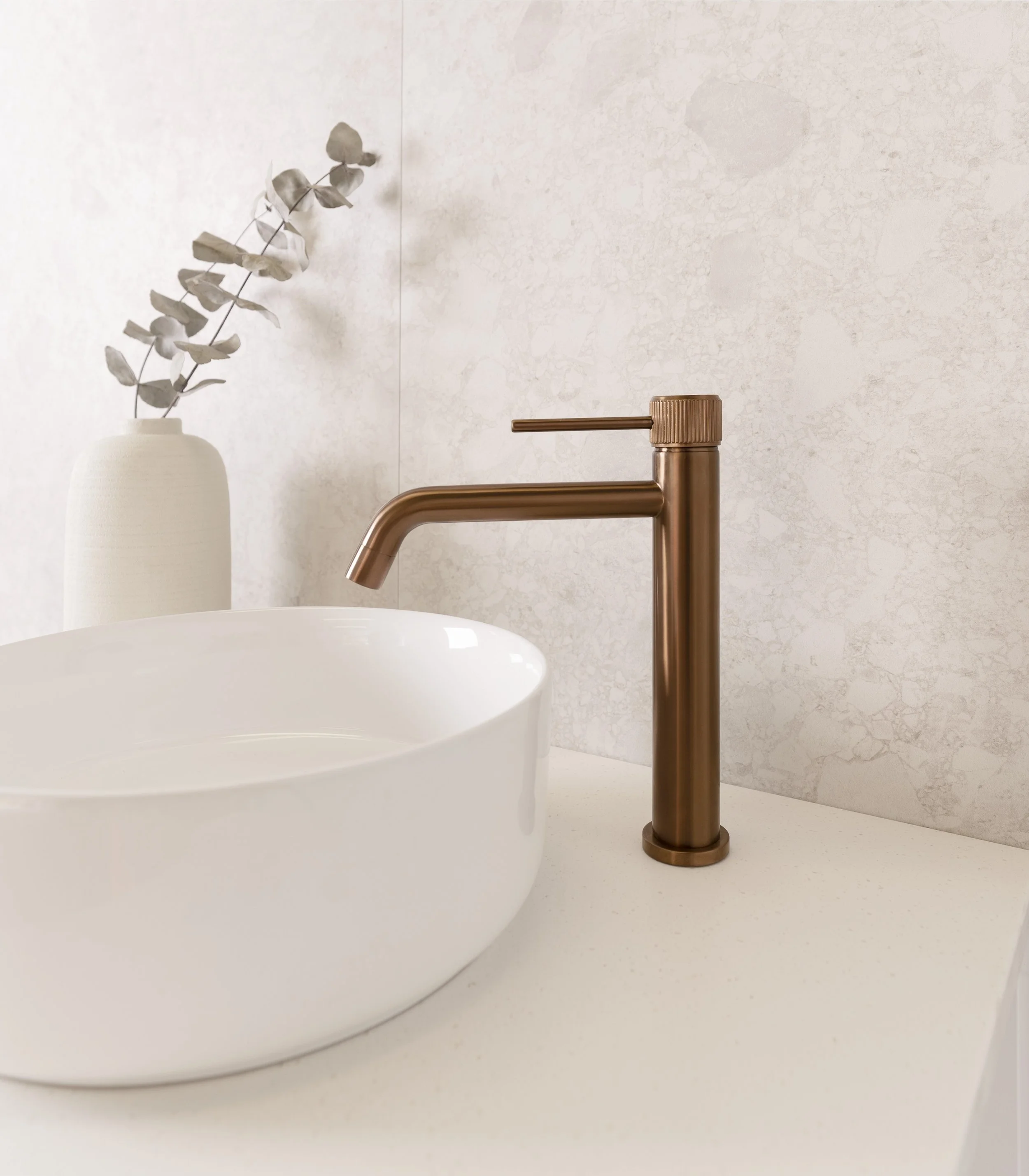 A minimalist bathroom sink area with a white vessel sink, bronze faucet, and a white vase with eucalyptus branches in the background.