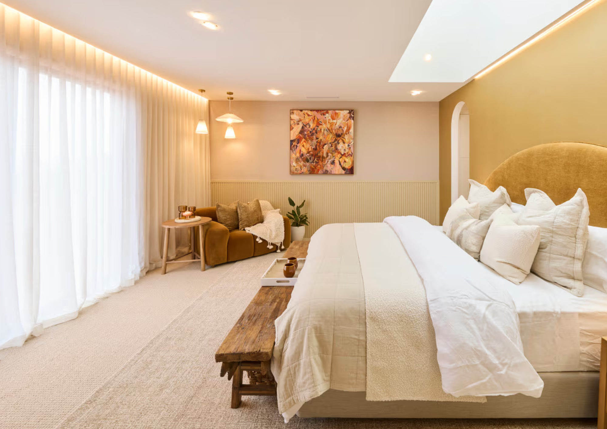 A cozy hotel bedroom with a large bed, white bedding, and multiple pillows. To the left, there is a small wooden bench at the foot of the bed. In the background, there is a sitting area with a mustard-colored sofa, a small wooden side table, and an abstract painting on the wall. The room has floor-to-ceiling curtains allowing natural light and warm ceiling lighting.