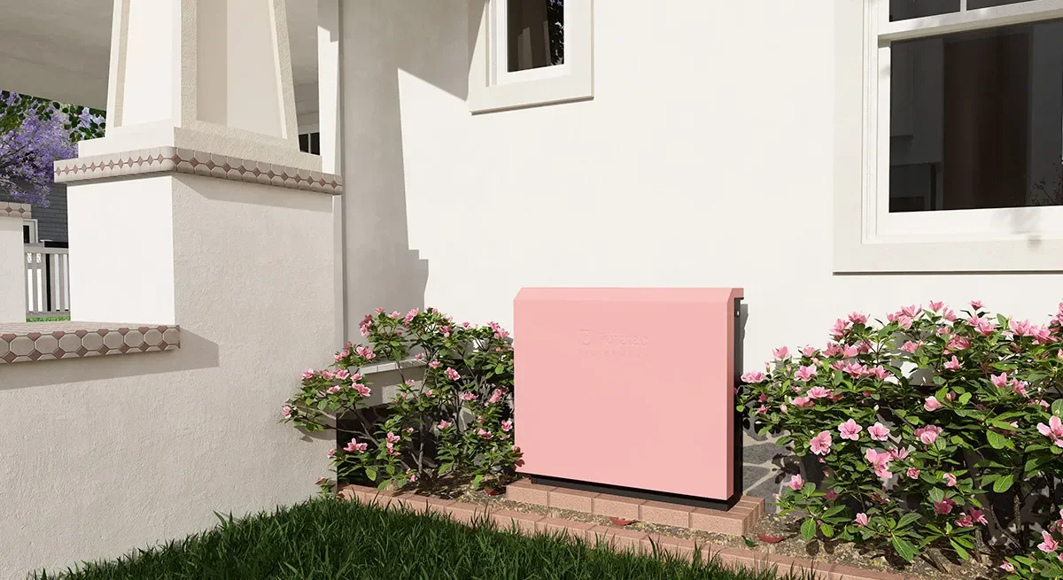 A pink mailbox on a brick base, surrounded by pink flowering bushes, outside a white house with a window and partial porch