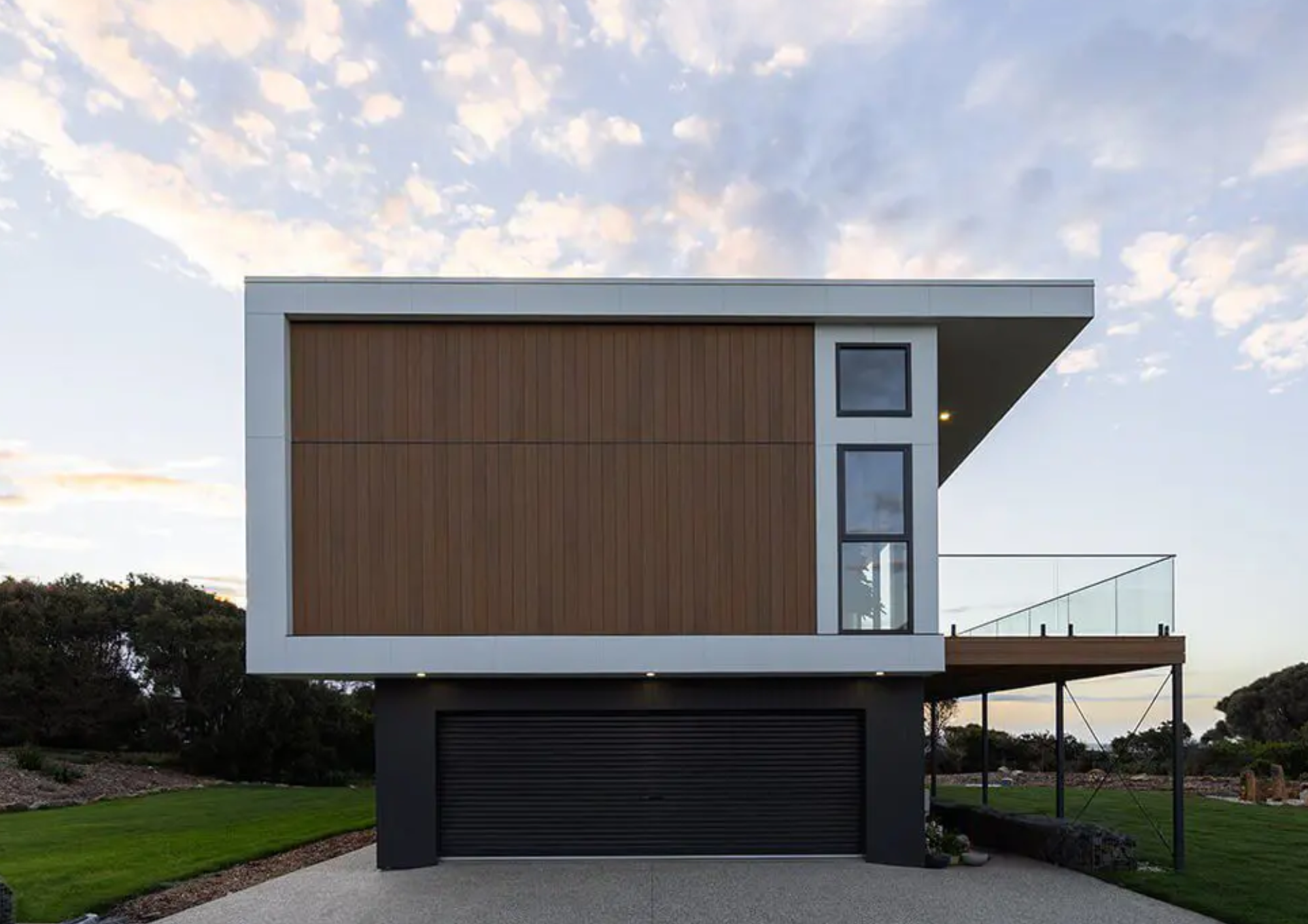 Modern, multi-story house with a flat roof, wood paneling, large windows, and a garage on the ground level, with a lawn and trees in the background.