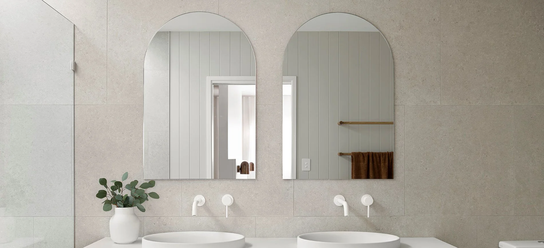 Bathroom vanity with two mirrors, white vessel sinks, eucalyptus plant in white pot, and brown towels hanging on wooden rods on a beige tiled wall.