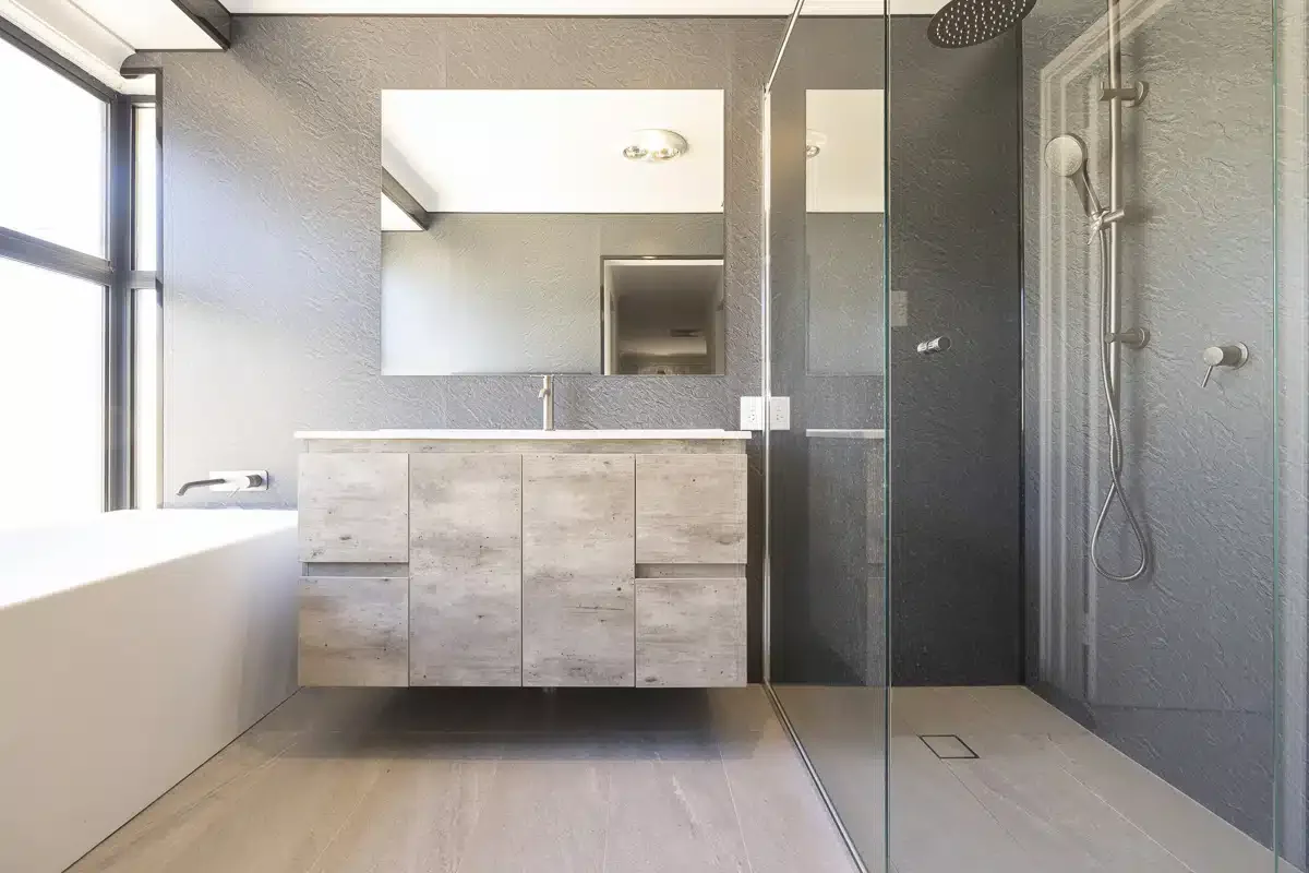 A modern bathroom with a large mirror, a wooden vanity with a sink, a window with natural light, and a walk-in shower with gray textured walls and a rain showerhead.