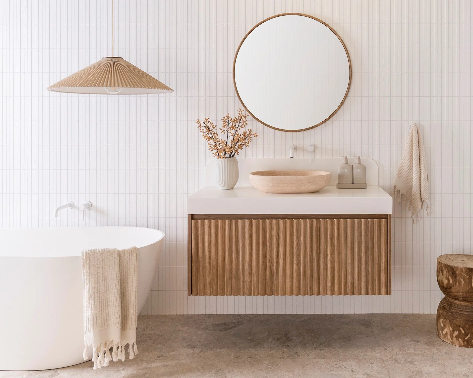 Modern bathroom with a white bathtub, a wooden vanity with a vessel sink, a large round mirror, a hanging pendant light, a vase with dried flowers, and a wooden stool.