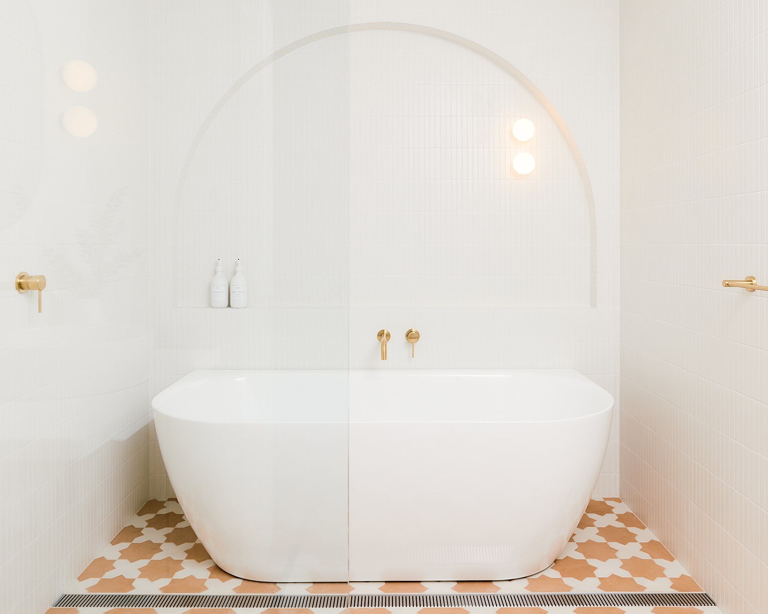 Modern white bathtub with gold fixtures, two bottles on a ledge behind, in a minimalistic bathroom with beige and white tiled walls and patterned floor.