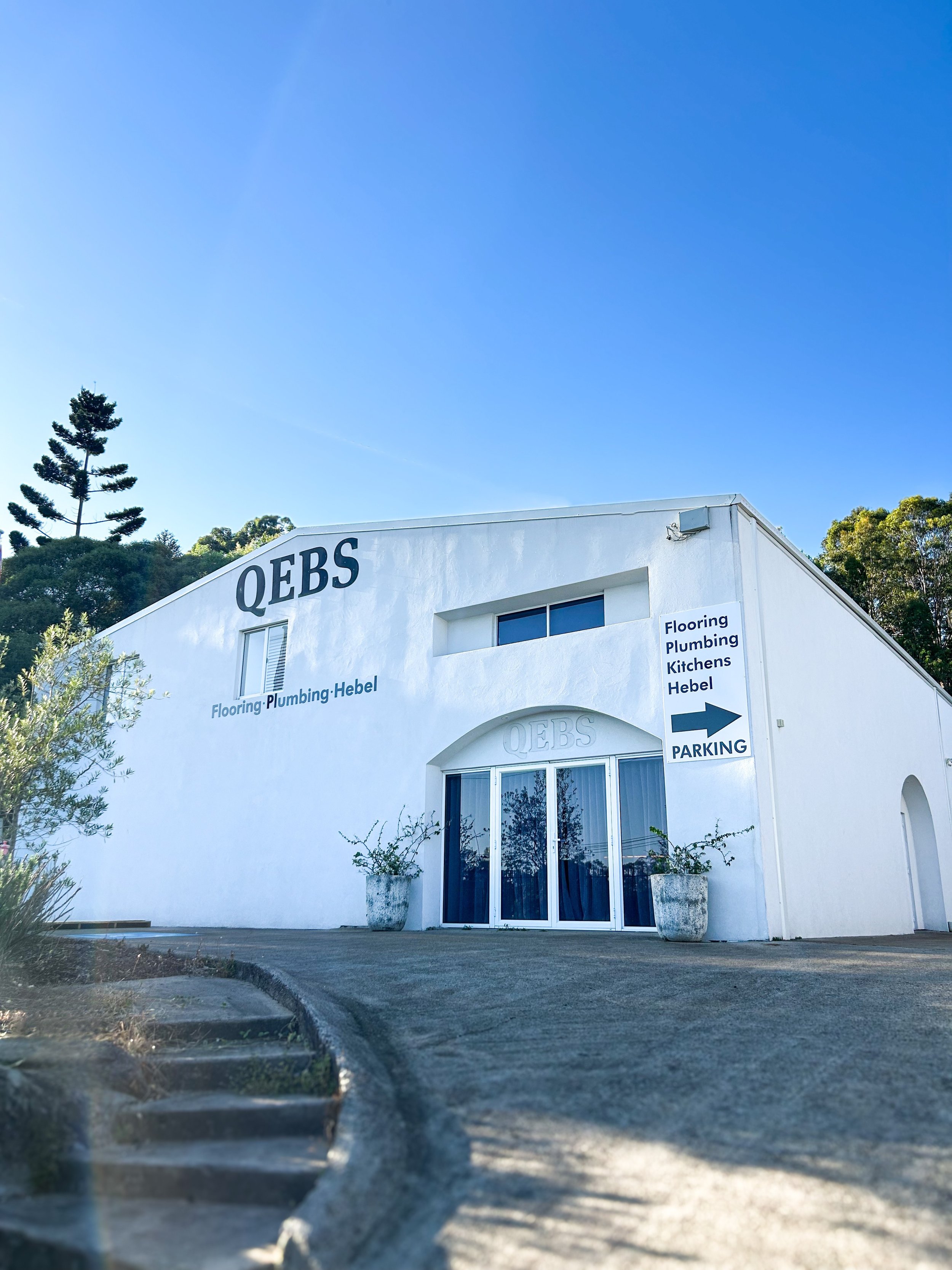 A white commercial building with the sign 'QEBs'. The building has an arched entrance with glass doors, potted plants on either side, and signs indicating services like flooring, plumbing, and kitchens. There is a parking sign and a directional arrow. The sky is clear and blue, with trees in the background.