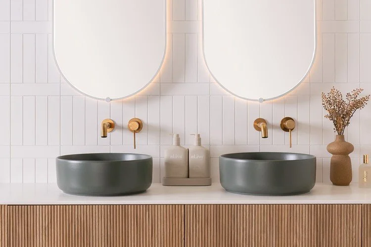 Double bathroom vanity with two oval gray sinks, two round brass faucets, oval backlit mirrors, a vase with dried flowers, and bottles of soap on a countertop with vertical wood paneling.