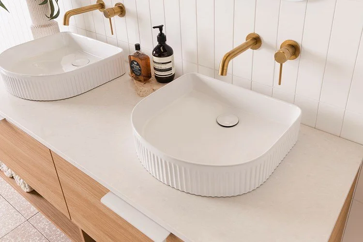 Double bathroom vanity with two white ceramic sinks, gold faucets, and soap dispensers on a white countertop in a modern bathroom.