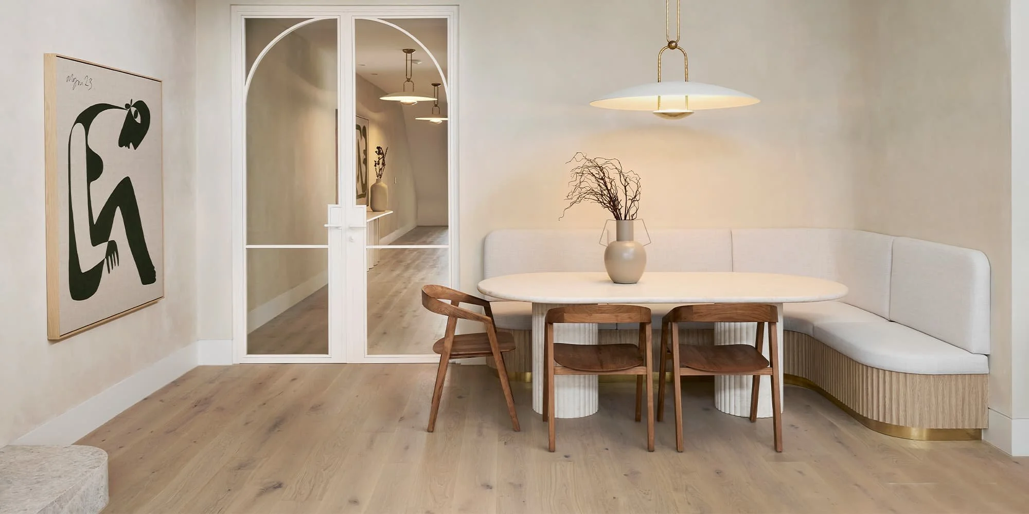 Minimalist dining area with a white curved banquette, a white oval table, and wooden chairs. A hanging ceiling light, beige walls, wooden flooring, a painting on the wall, and a door with glass panes leading to a hallway with matching lighting fixtur
