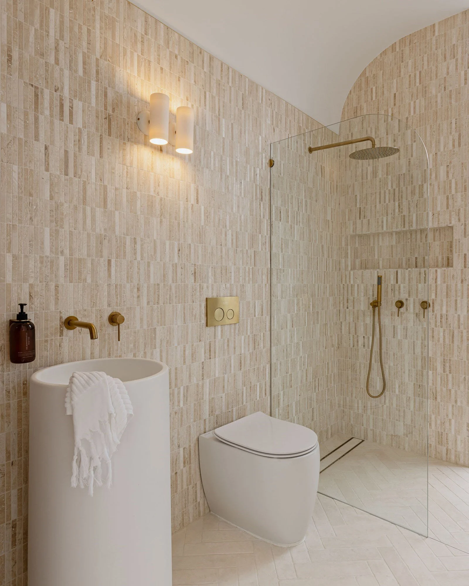 Modern bathroom with beige tiled walls and a walk-in shower with a glass partition, a wall-mounted showerhead, and a handheld shower. Features a white toilet and a white freestanding sink with a black soap dispenser and a white towel hanging on the s