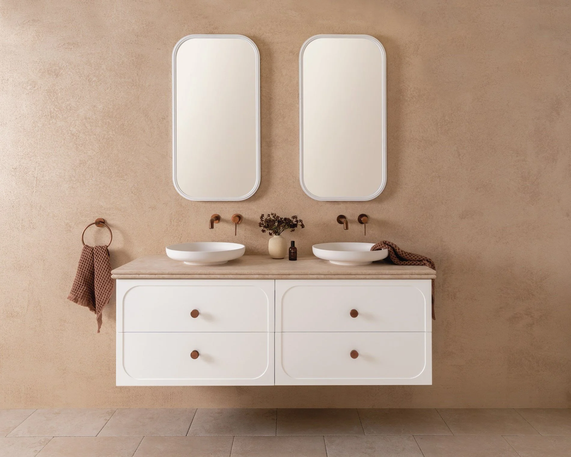 A modern double vanity bathroom with two oval vessel sinks, two oval mirrors, brown soap dispensers, and brown towels hanging on a wall-mounted towel ring and draped over the sink. The vanity has four drawers with simple round knobs and a beige count