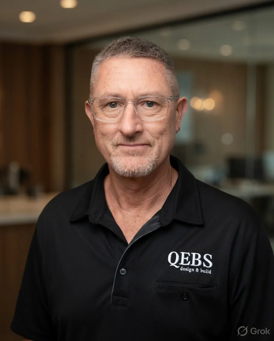 A middle-aged man with short gray hair, glasses, and a goatee, wears a black polo shirt with the QEB logo and stands in an indoor setting with blurred background.