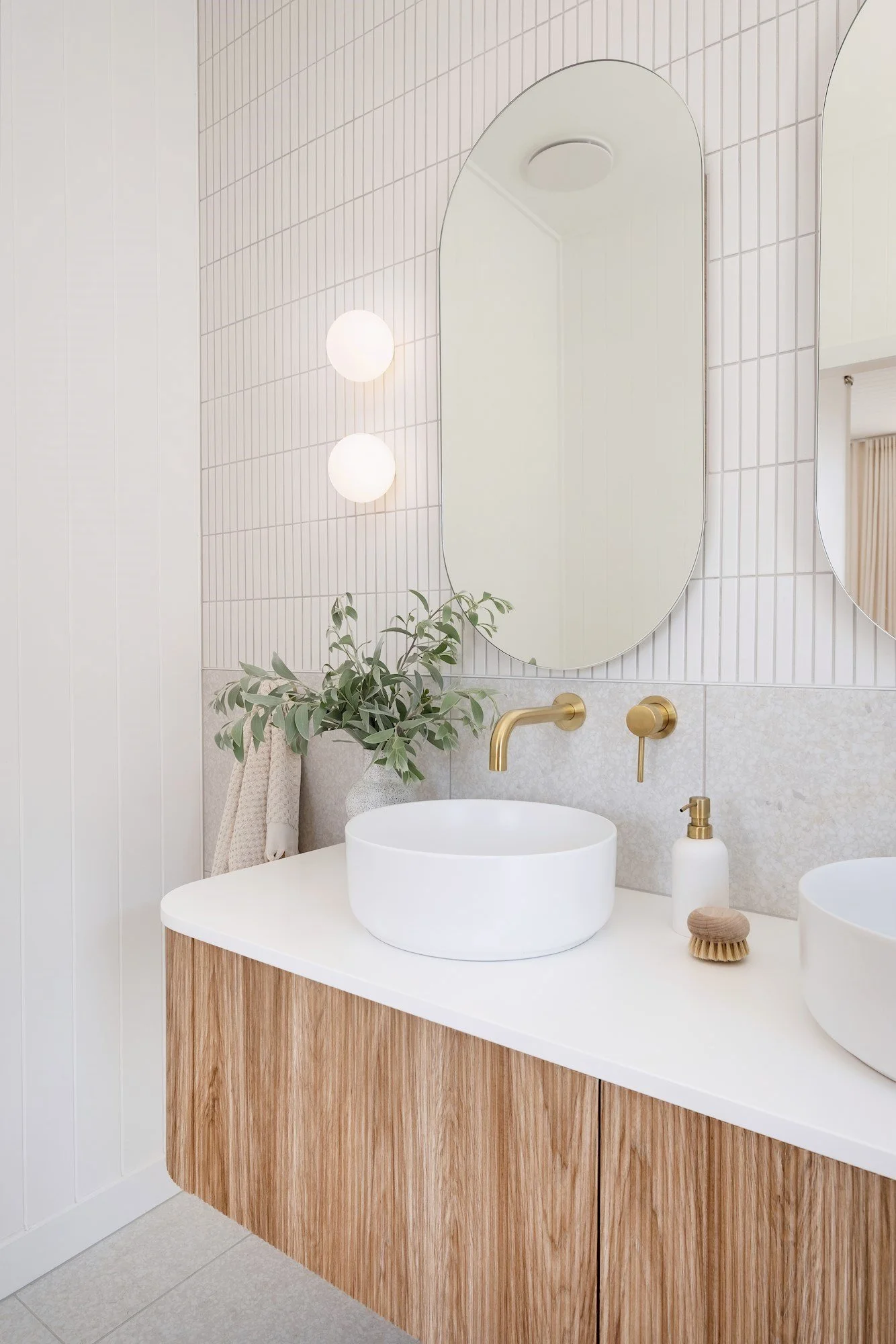Modern bathroom with white vessel sinks, gold fixtures, and large mirrors, decorated with a plant, soap dispenser, and grooming brush.