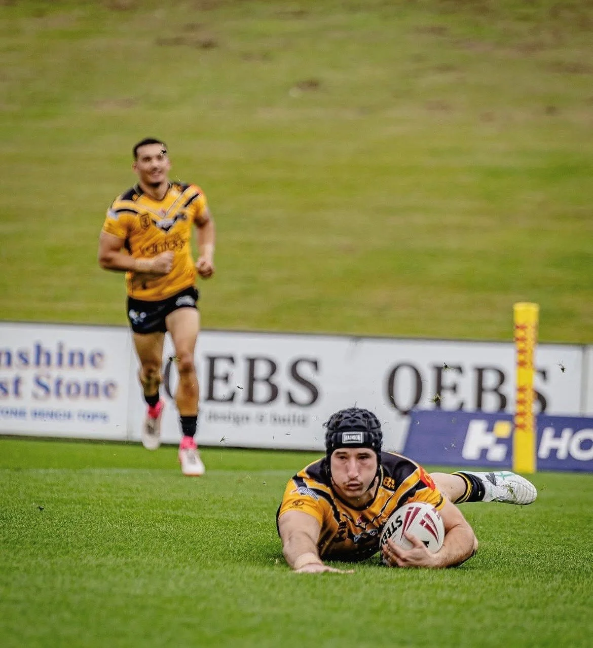 Two rugby players in yellow and black jerseys, one diving on the field with a ball, the other running in the background on a grassy field.