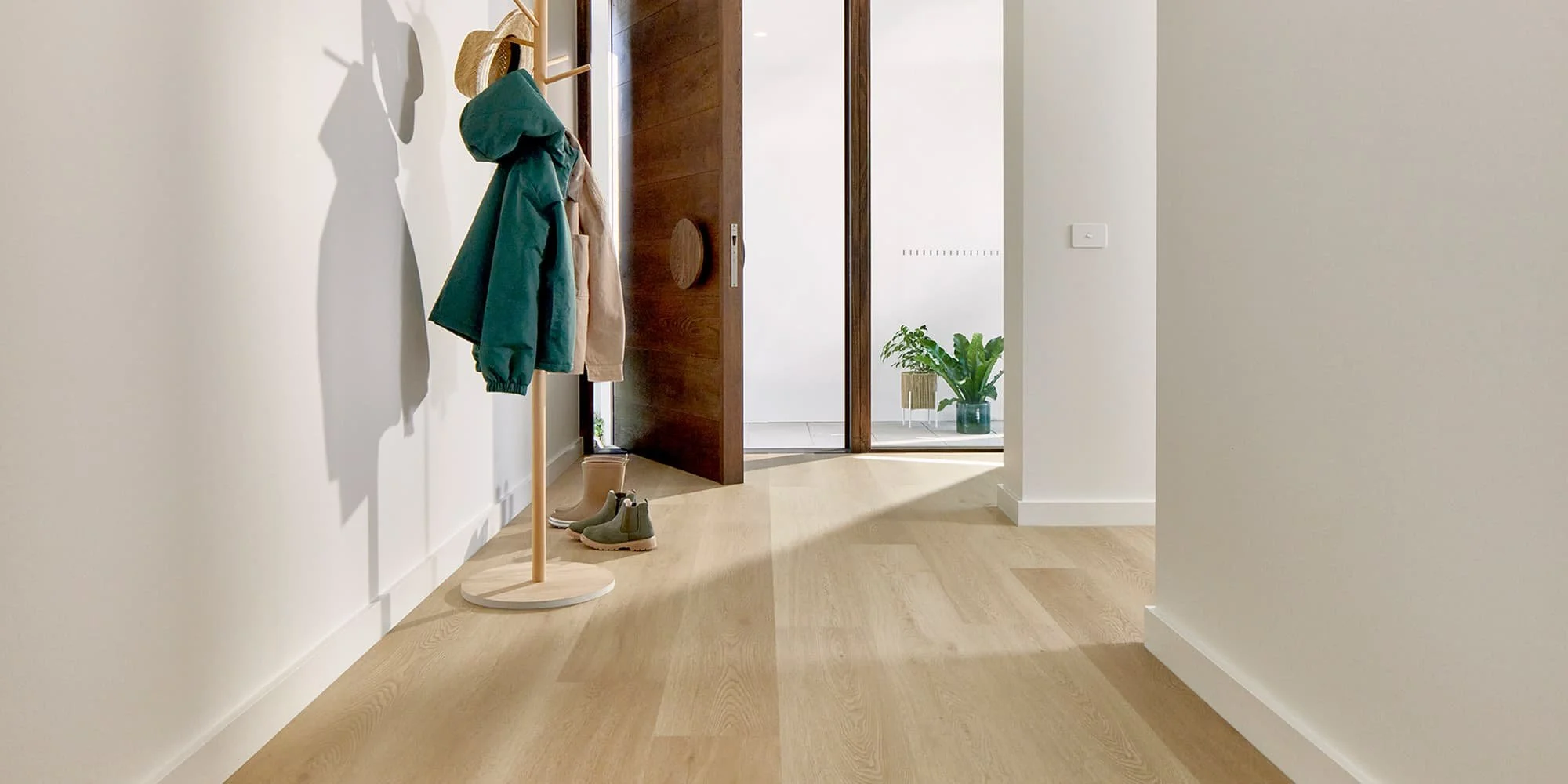 Entryway with a wooden coat rack holding jackets and hats, beige boots and slip-on shoes on the floor, open wooden door, and a potted plant in the light-filled room beyond.