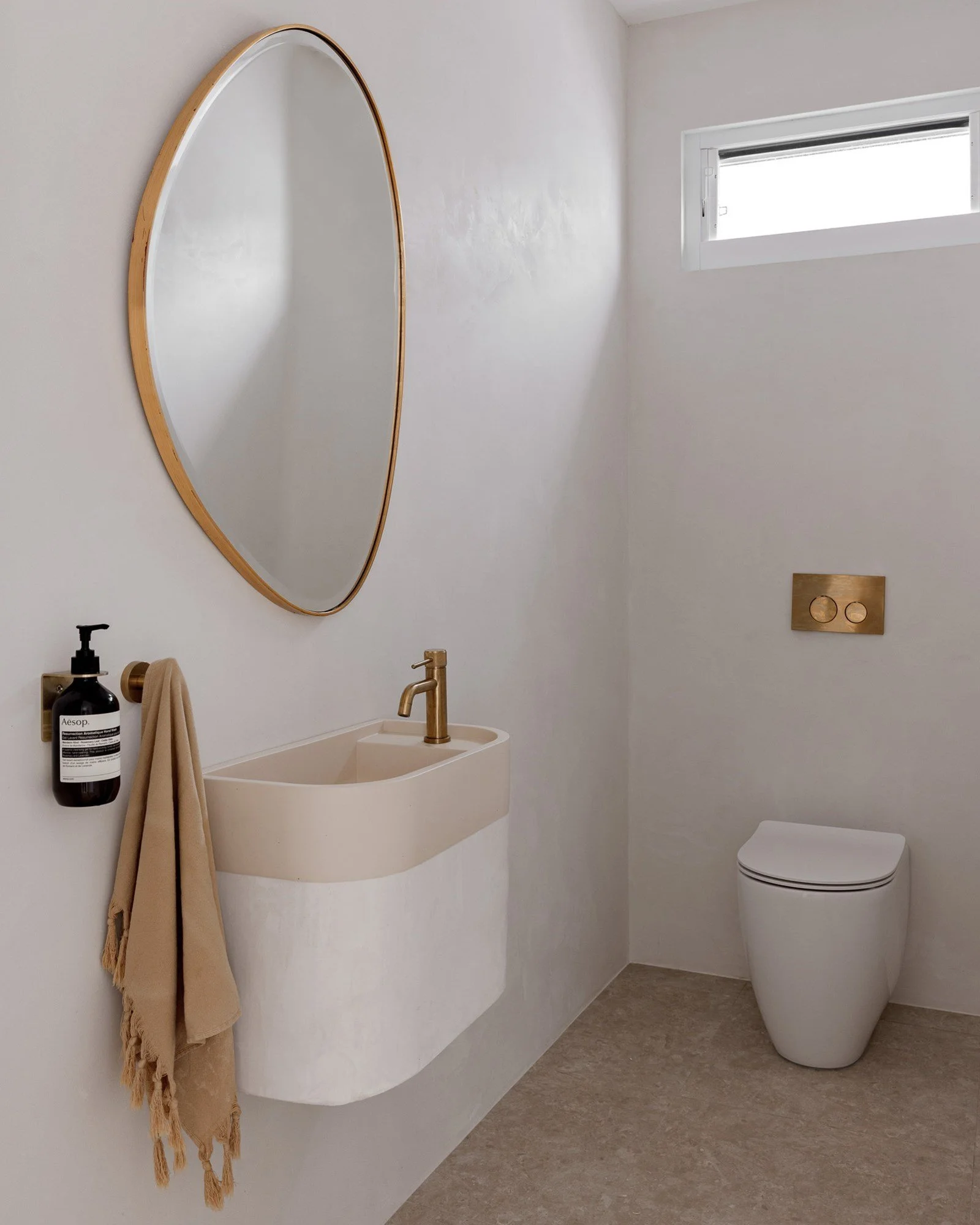 Modern bathroom with a white wall-mounted sink, gold faucet, oval mirror with wooden frame, black soap dispenser, beige towel, and a white toilet with gold flush panel, small window letting in natural light.