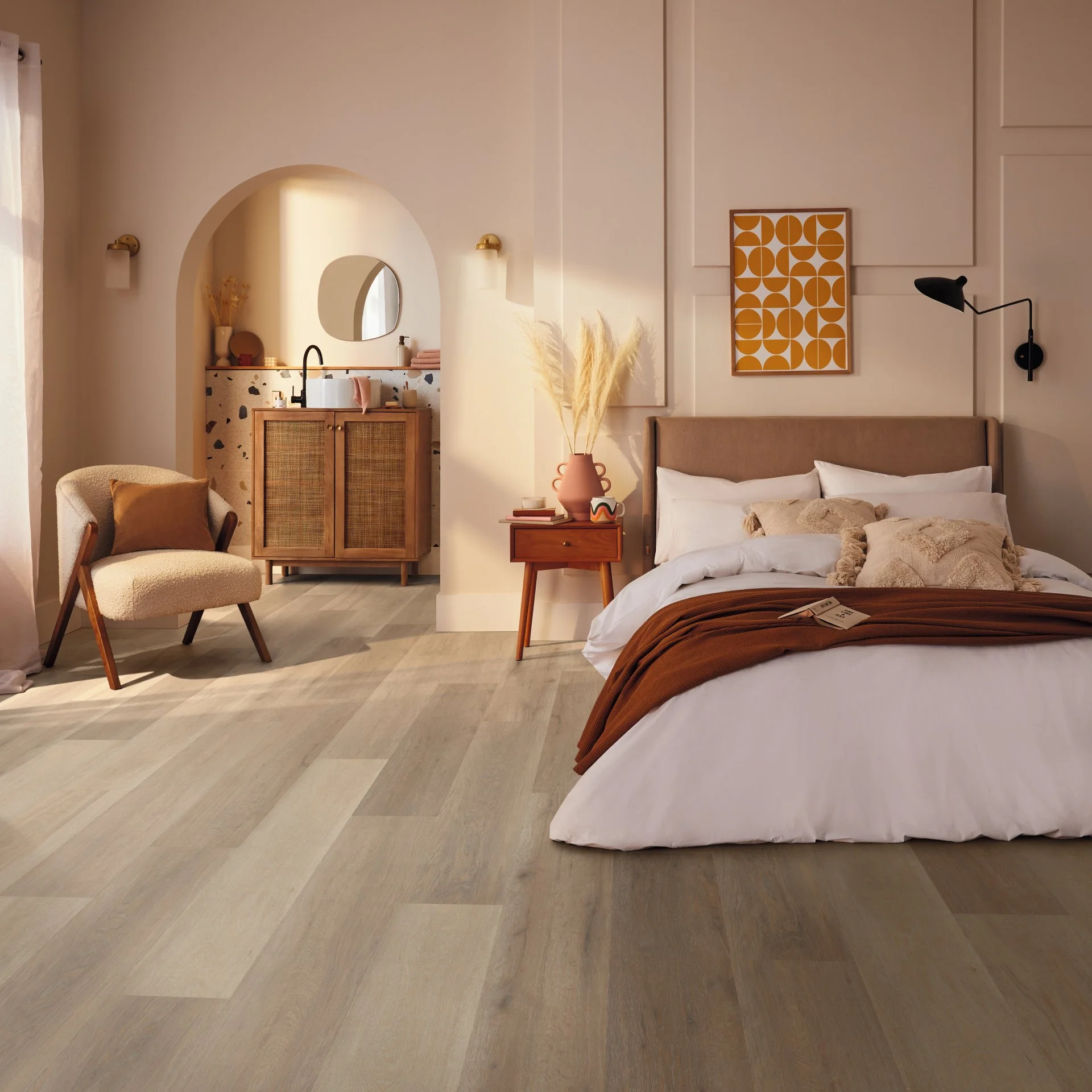 A cozy bedroom with a neatly made bed, a tan headboard, and neutral-colored pillows. There is a small wooden side table with a pink vase, pampas grass, and books. To the left, a cream armchair with a cushion is illuminated by natural light. Behind th