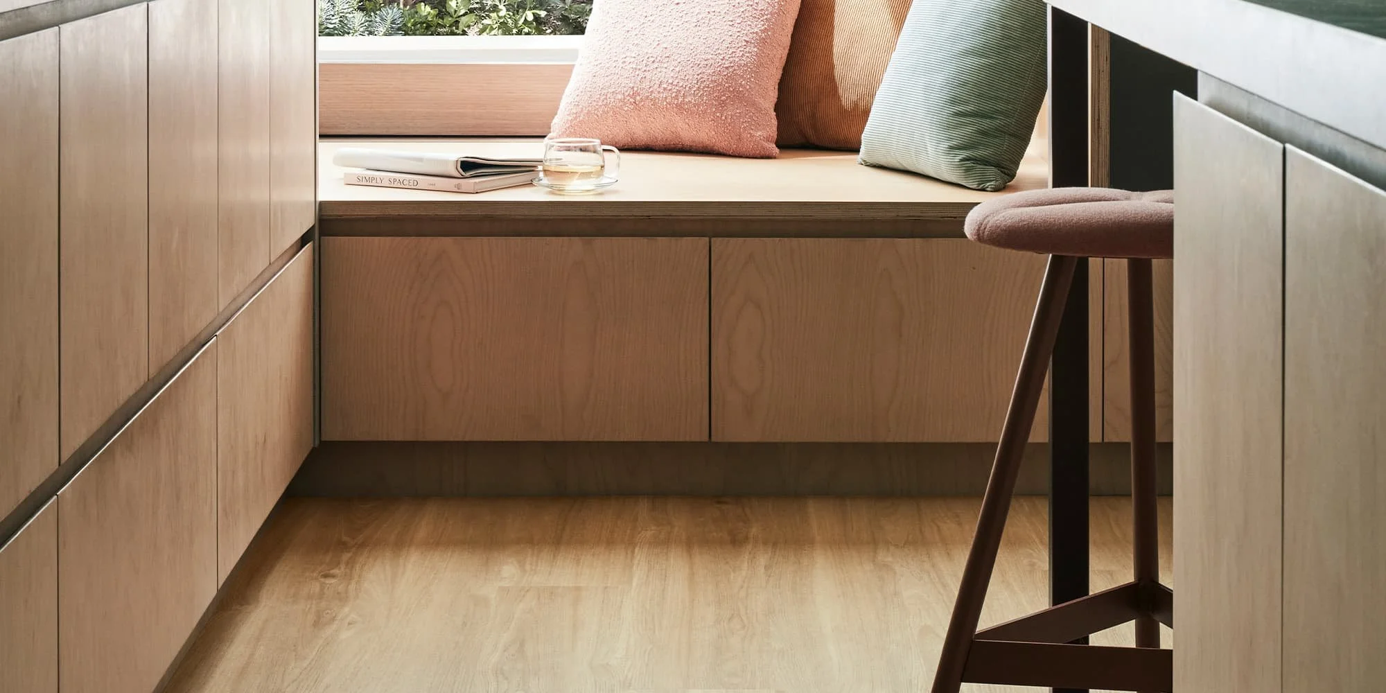 A cozy kitchen nook with a built-in wooden bench under a large window, decorated with pastel-colored cushions and a small glass of water on a book titled 'Simply Spaces'.