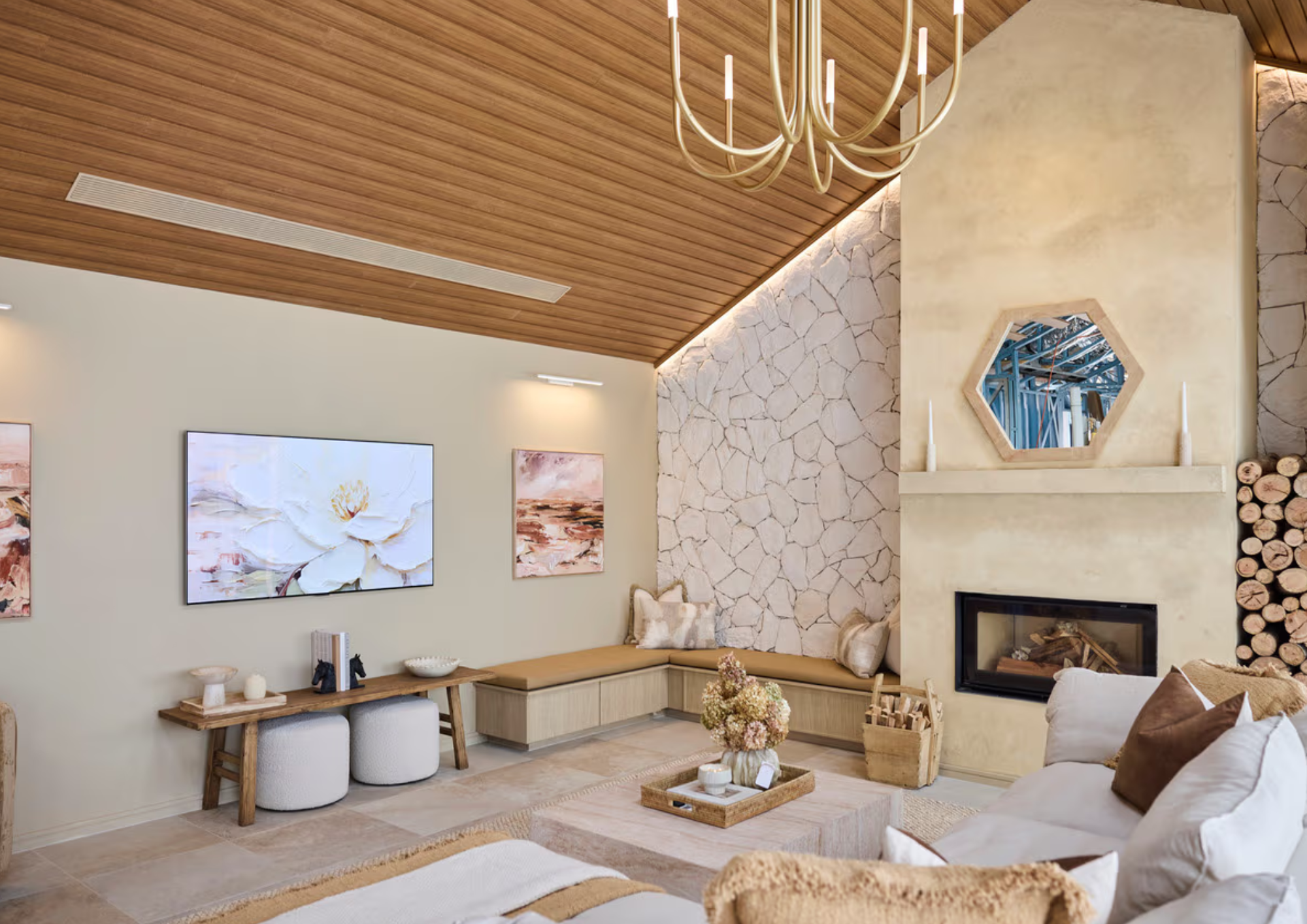 Living room with cream-colored sofa, fireplace, stone wall, wooden ceiling, chandelier, artwork, and a window reflecting a blue roof structure.