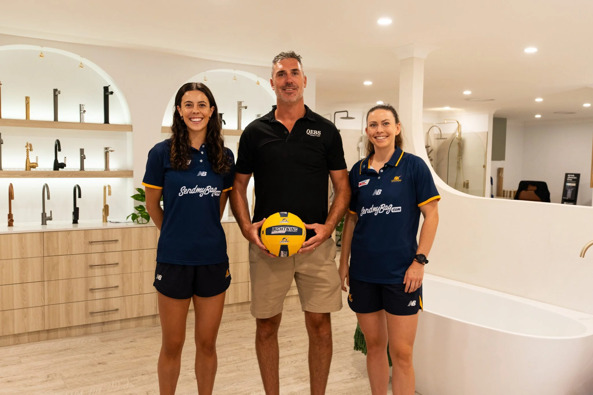 Three people standing in a room with a variety of kitchen faucets on shelves behind them. The person in the center is holding a yellow volleyball, and all three are smiling.
