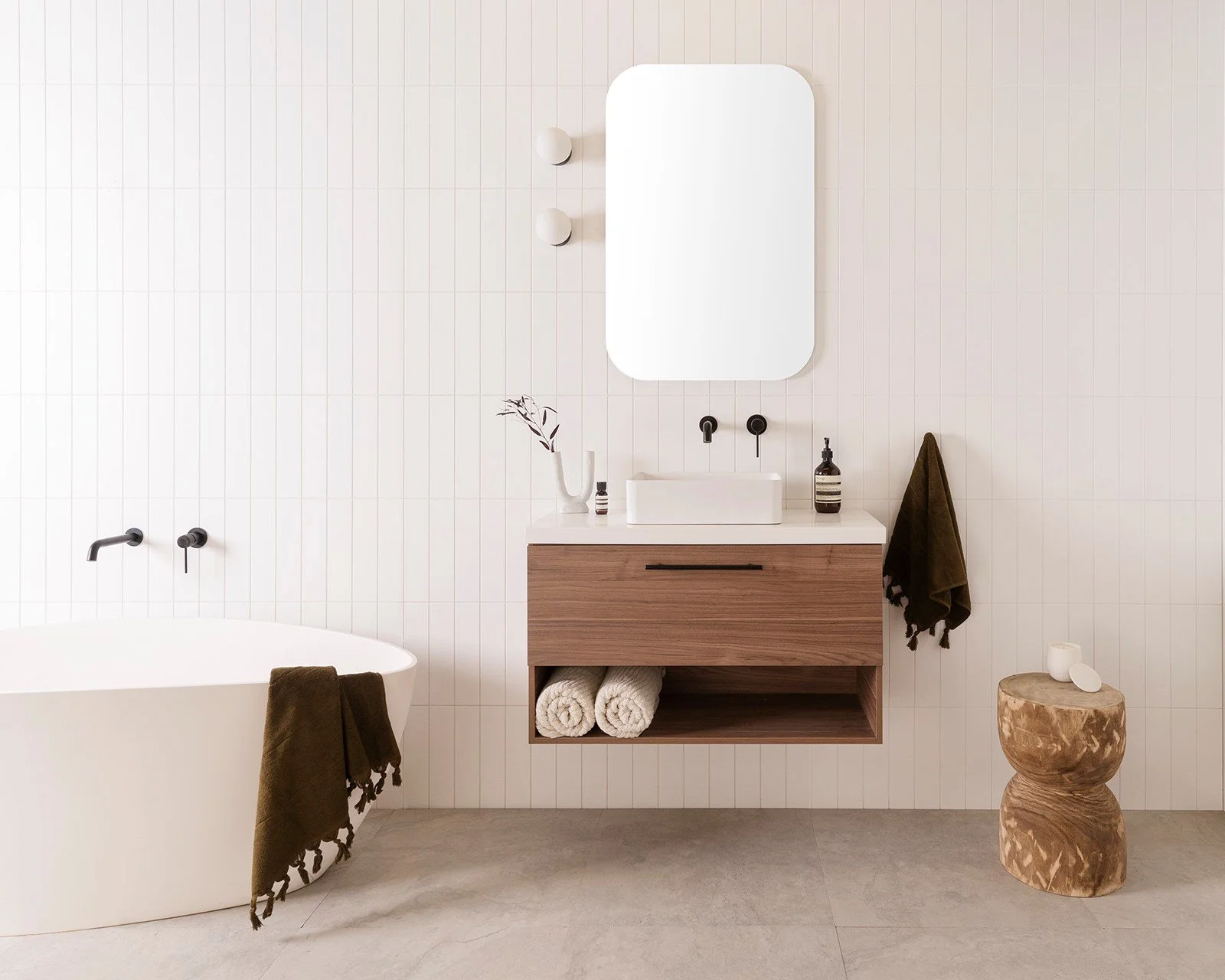 Modern bathroom with a white bathtub, a wooden vanity with a white sink, a tall mirror, and minimal decor including towels, a small vase, and soap.
