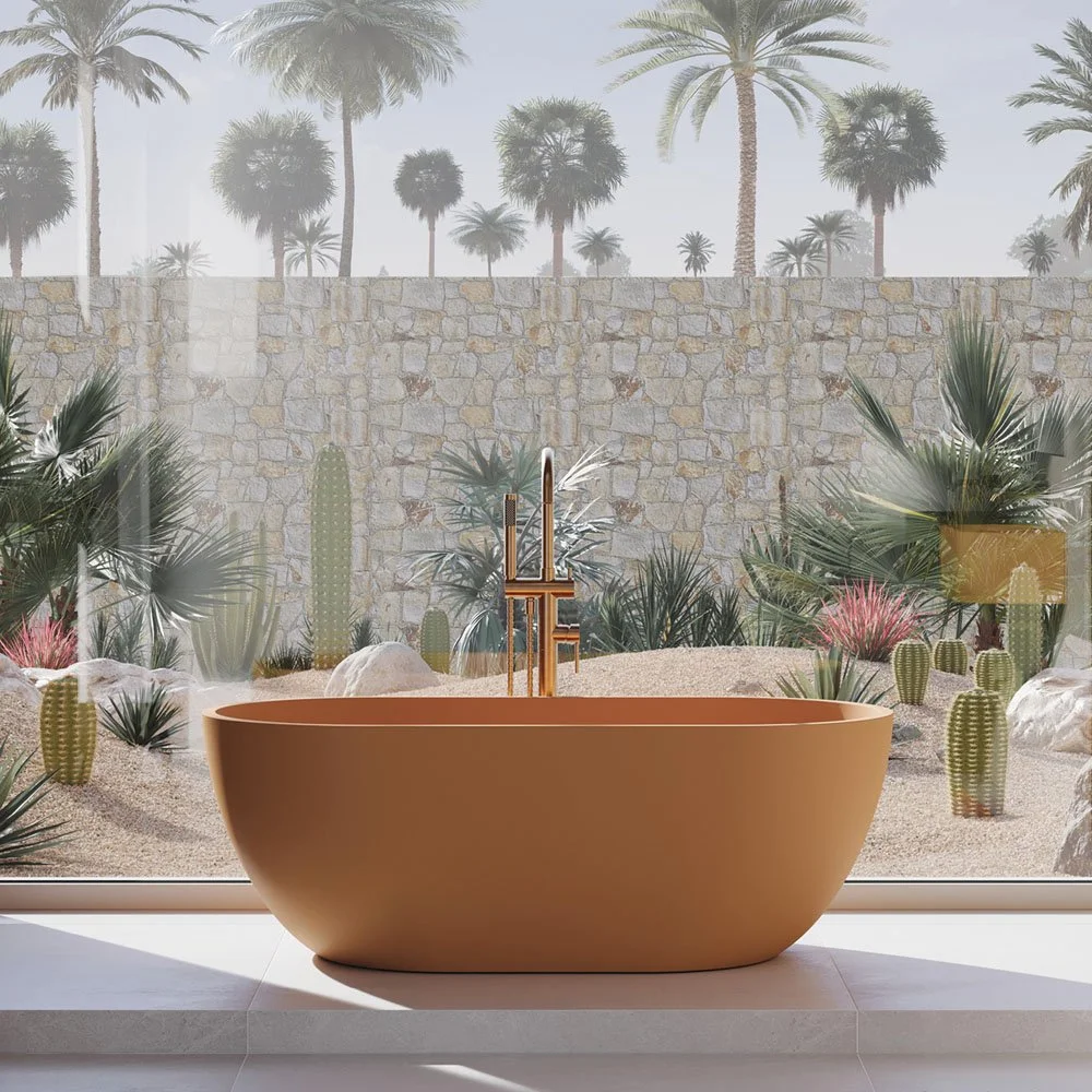 A tan, freestanding bathtub with gold fixtures is positioned in front of a large window revealing a desert landscape with cacti, palm trees, and a stone wall.