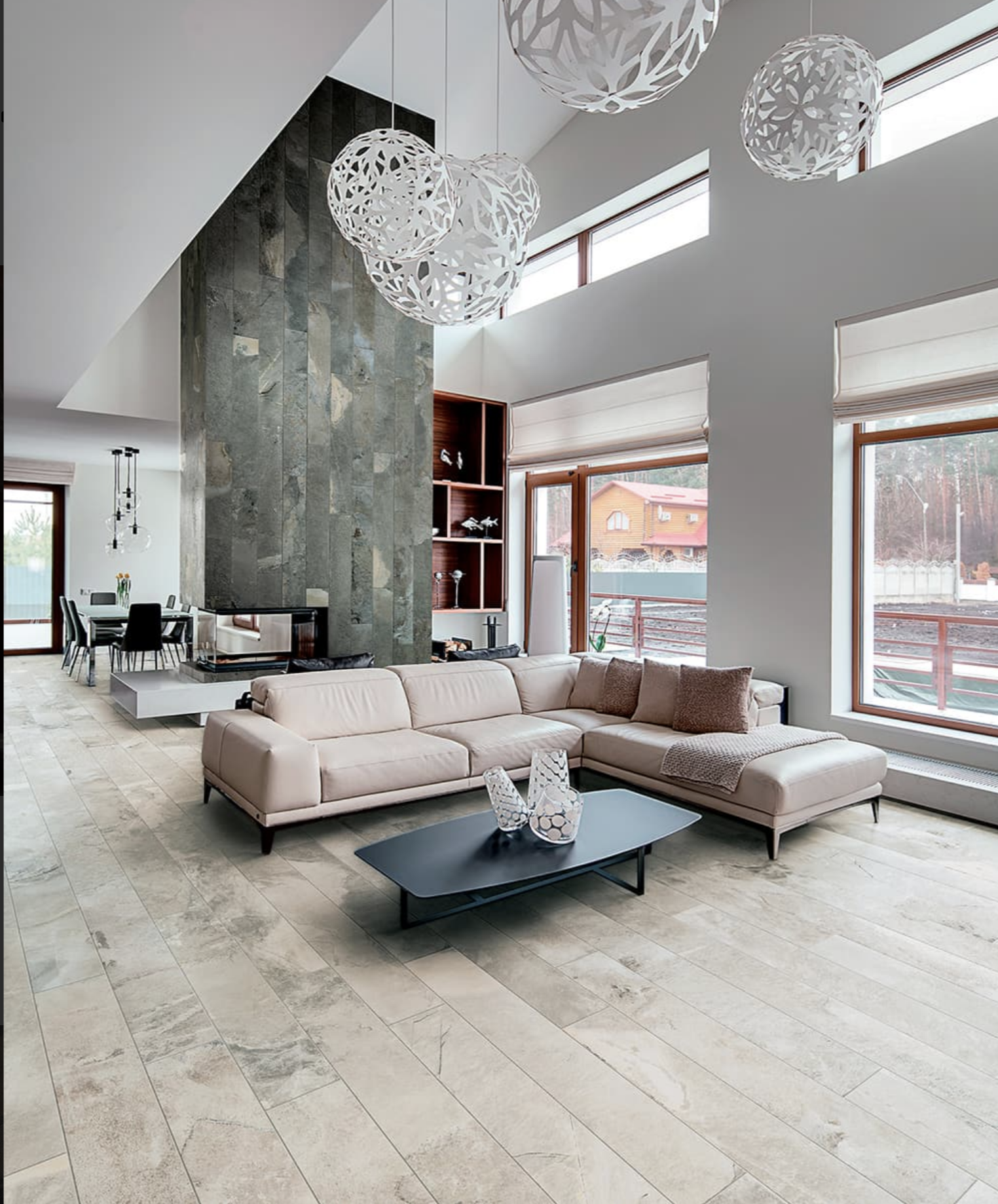Modern living room with a large L-shaped beige sofa, black coffee table, and decorative vases. Bright space with large windows, hanging white geometric pendant lights, and a stone fireplace.