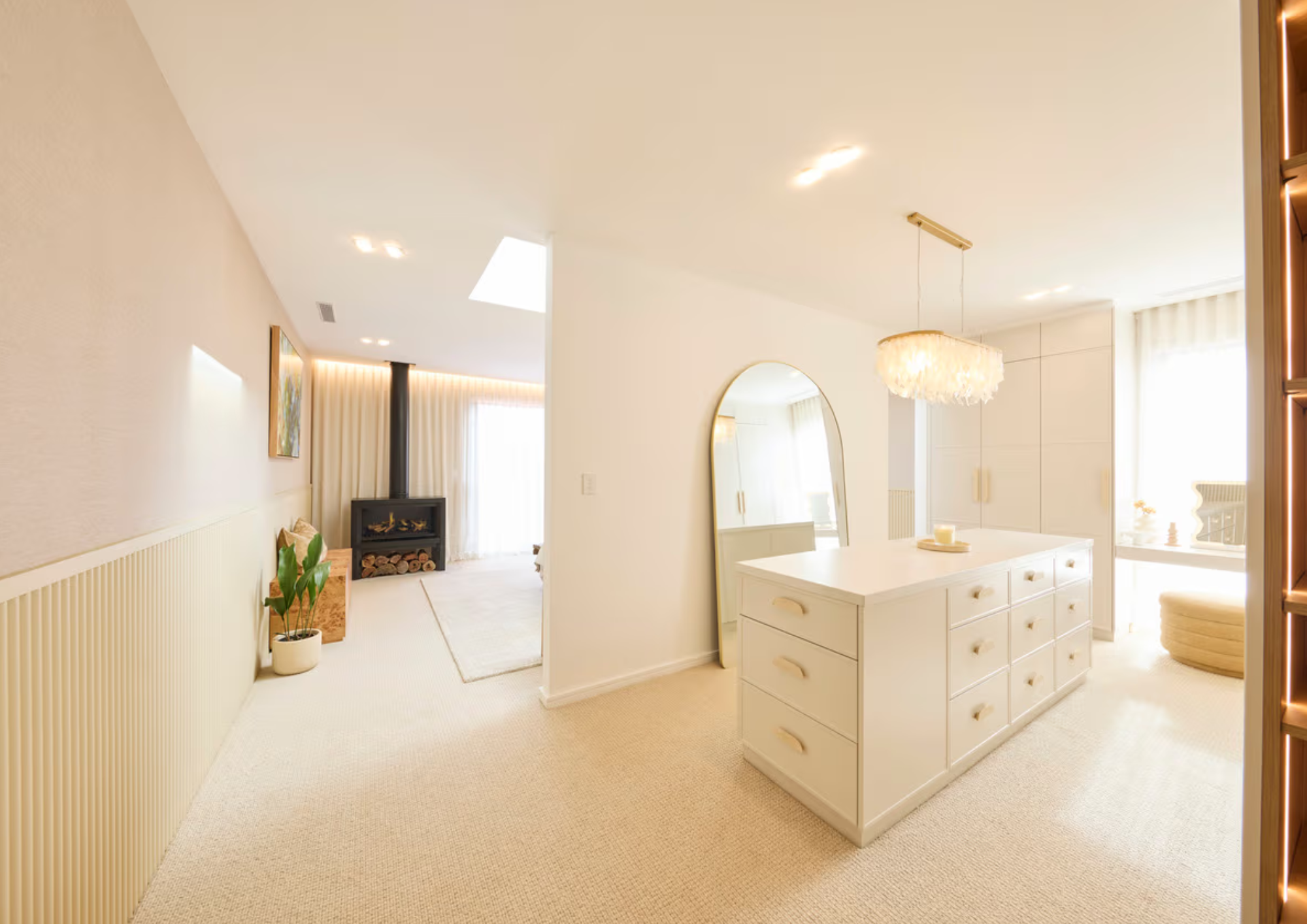 Bright and airy living and dining area with white walls, beige carpet, a white kitchen island, large window with curtains, a round mirror, chandelier, and a corner with a wood stove and plants.