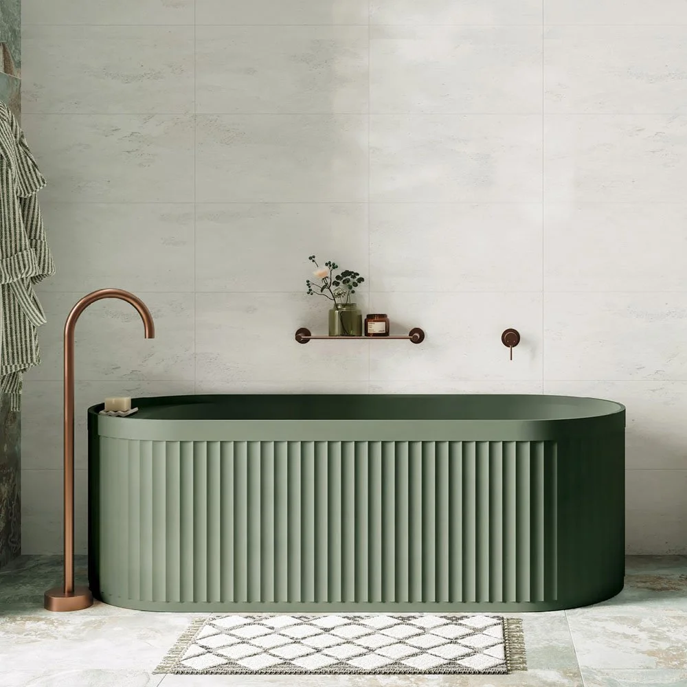 A modern bathroom with a green freestanding bathtub that has a vertically ribbed exterior. The bathtub is next to a tall copper-colored floor faucet. A small white rug with a diamond pattern is on the floor in front of it. The wall behind has large l