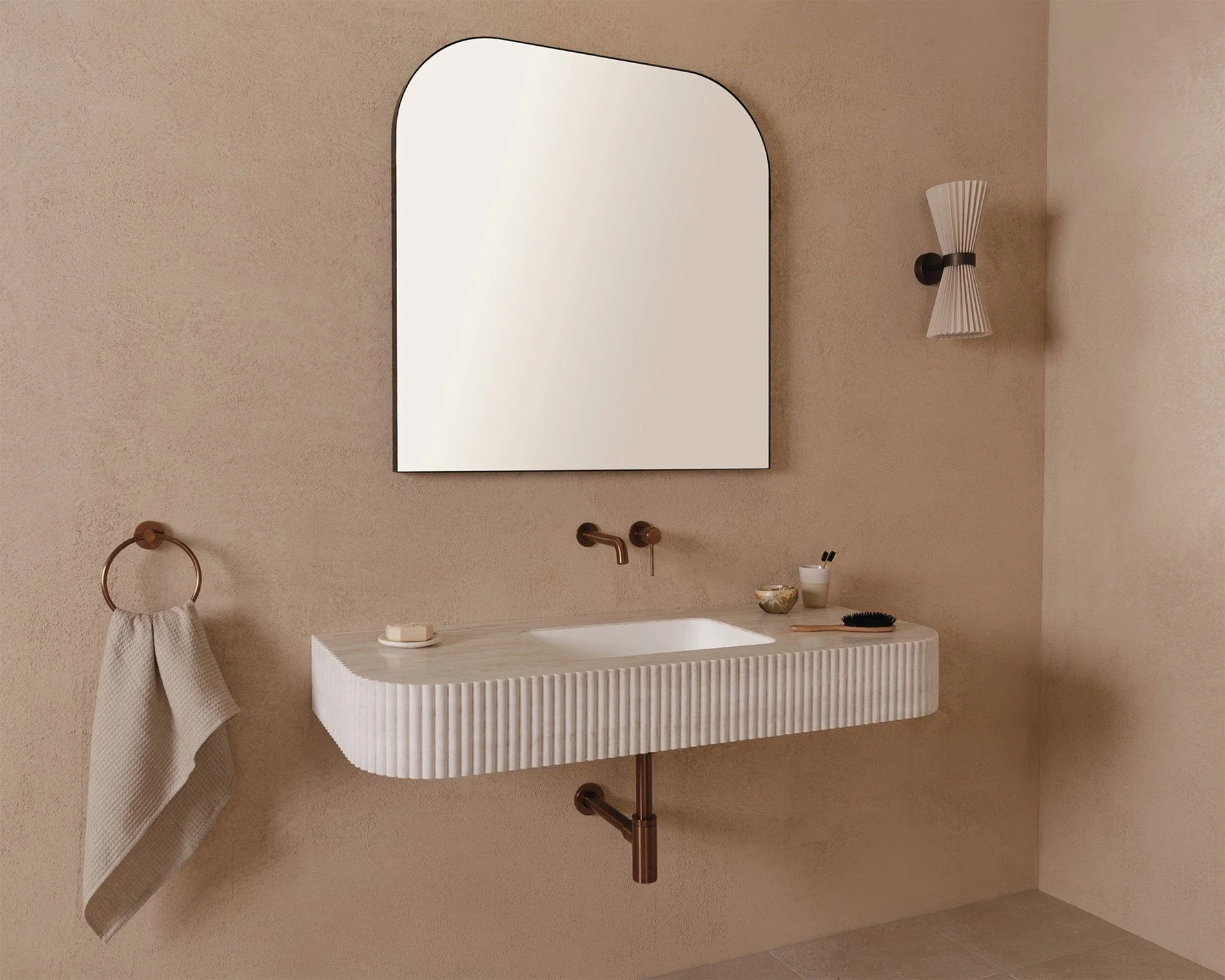 Modern beige bathroom with a wall-mounted sink, a large arched mirror, a wall-mounted light fixture, a hand towel on a ring, and small personal items on the sink