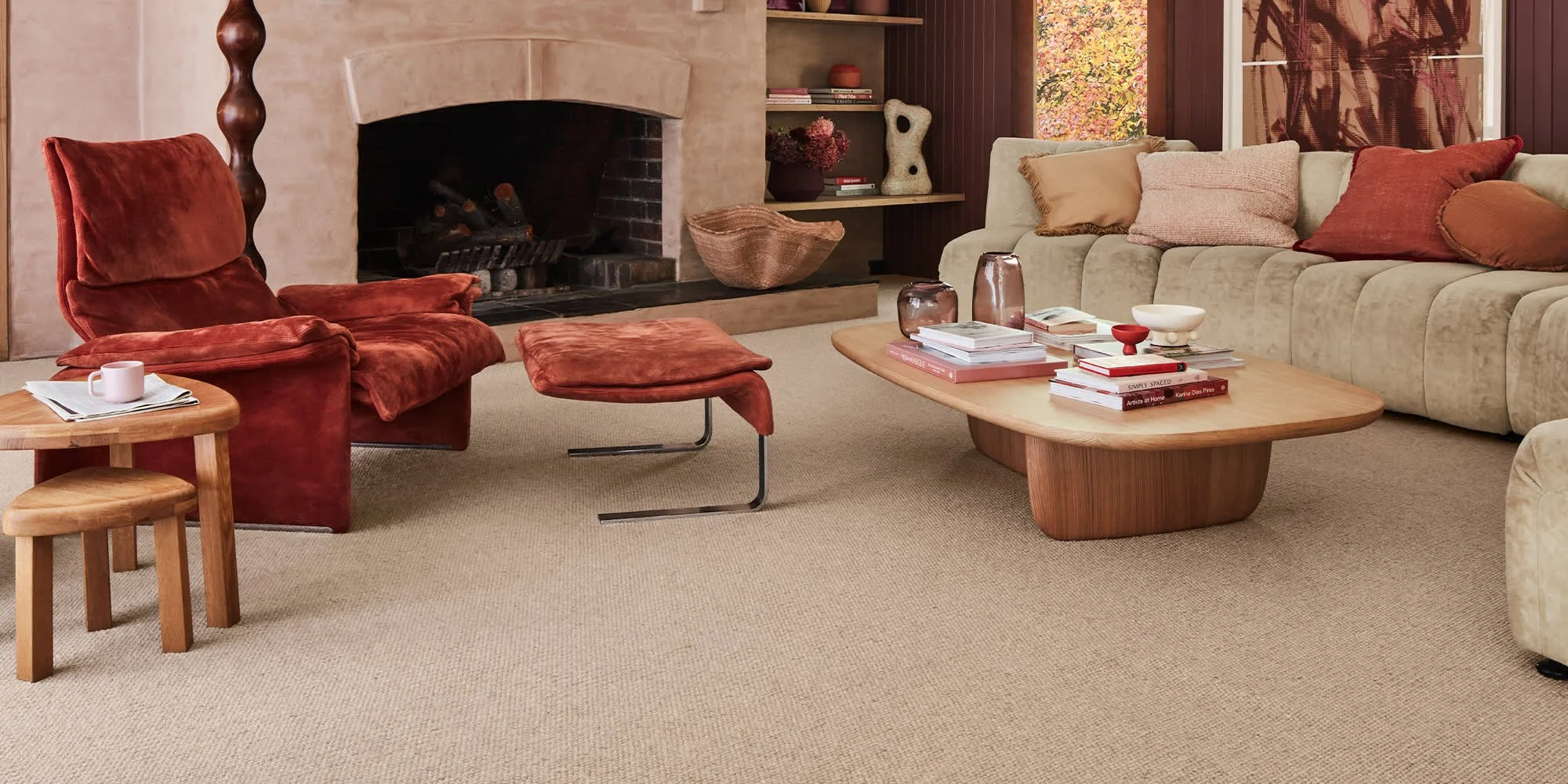 Living room with a beige carpet, red velvet armchair with a matching ottoman, beige sectional sofa with multiple cushions, wooden coffee table with books and decorative objects, small wooden side table with a cup, fireplace, and built-in shelves fill