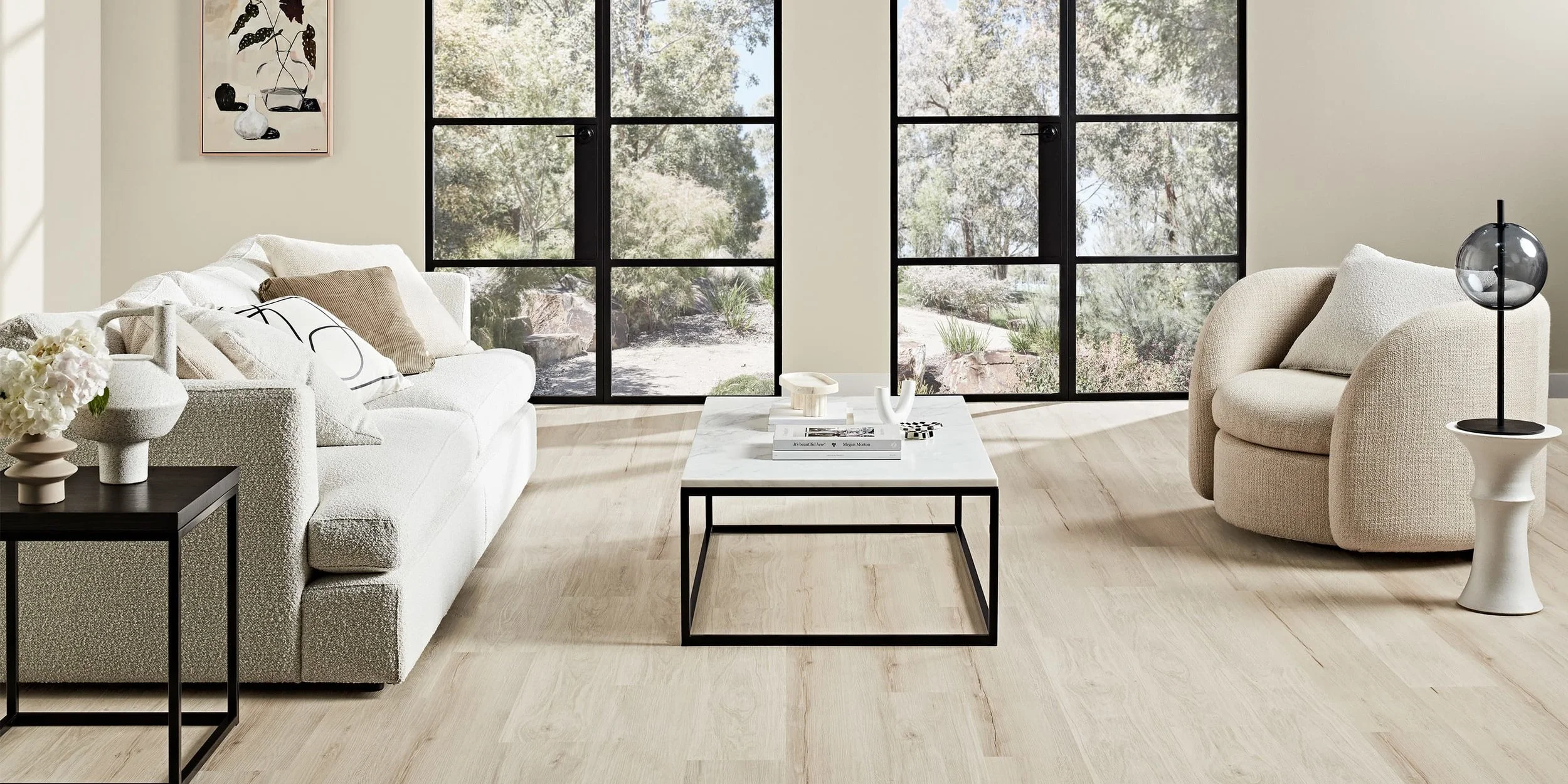 Modern living room with large glass windows, light-colored sofa, armchair, black coffee table, side table with flowers, minimalist decor, and natural light.