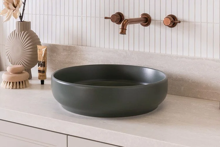A modern bathroom countertop with a dark gray round vessel sink, a beige decorative vase, and a small wooden brush and container.