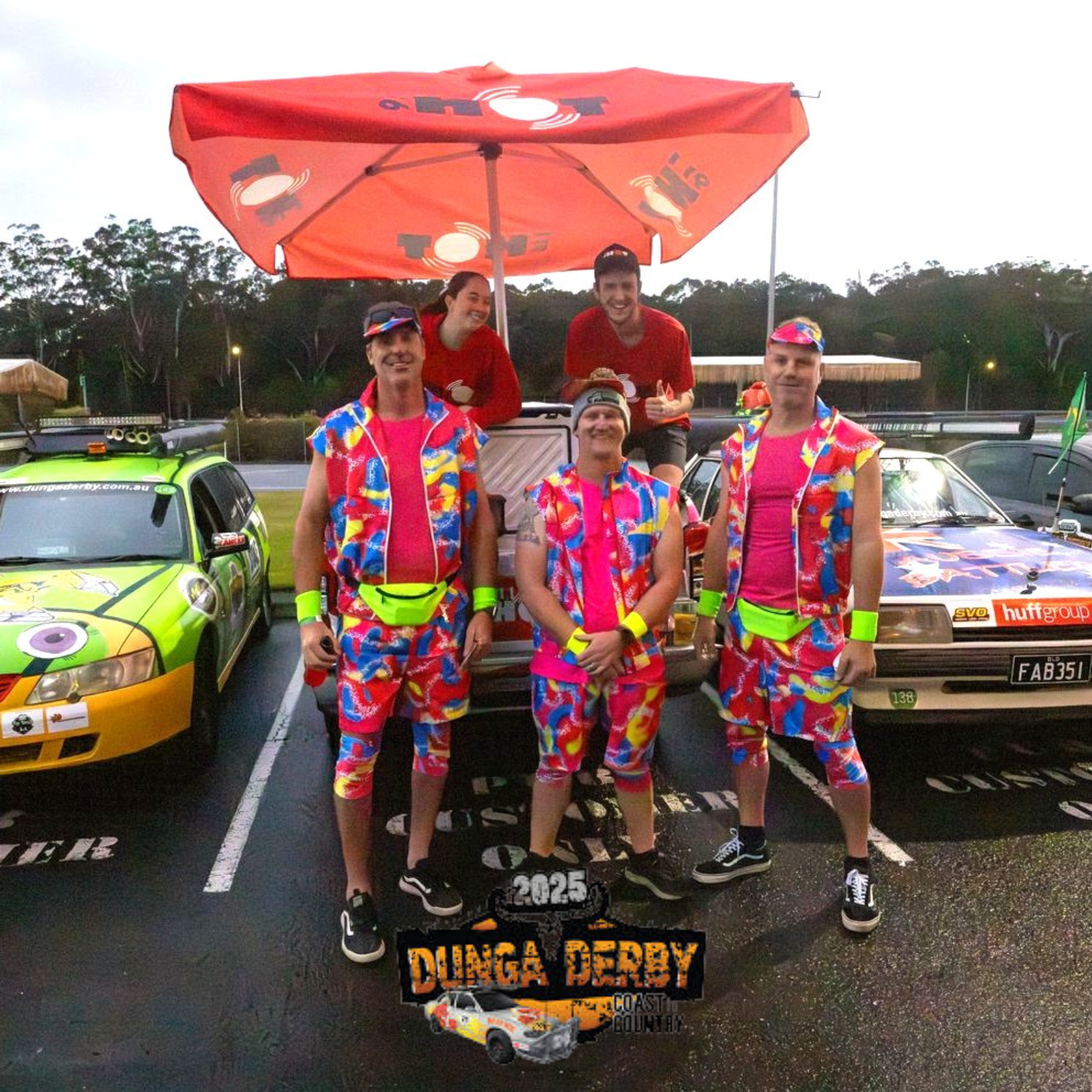Group of five people dressed in colorful, matching outfits outside in a parking lot during a car rally event, with two race cars behind them, and a large orange umbrella overhead. They are smiling and posing for the photo.