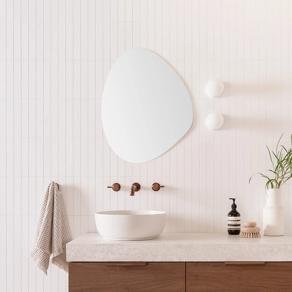 Modern bathroom with a white vessel sink, bronze wall-mounted faucet, oval mirror, two white wall sconces, a beige towel on a hook, black soap dispenser, a wooden brush, and a white vase with a green plant on a wooden vanity.