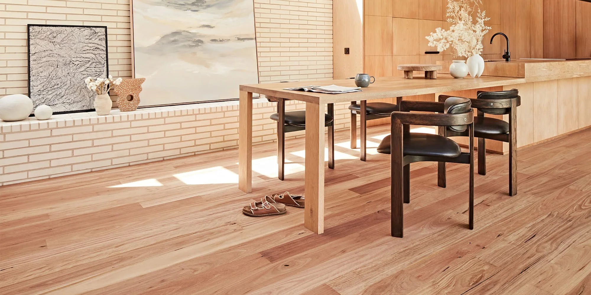 A minimalist kitchen and dining area with a wooden table, black chairs, decorative vases, artwork, and a pair of sandals on the wooden floor.