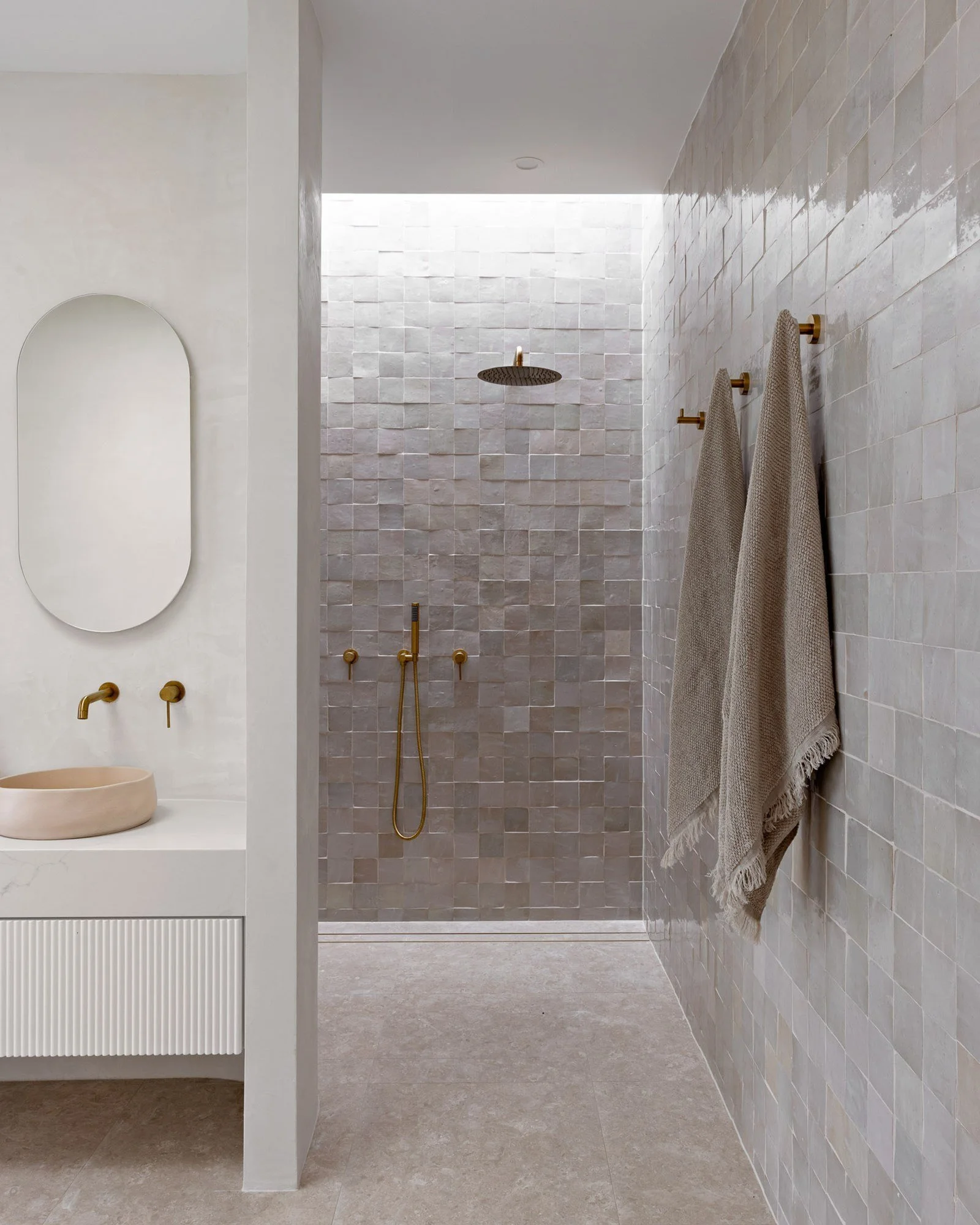 Modern bathroom with beige tiled walls, walk-in shower with rainfall showerhead, and beige towels hanging on wall hooks.