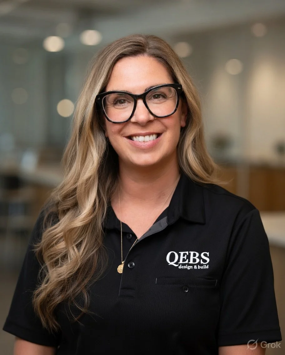 A woman with long, wavy blonde hair and black-rimmed glasses smiling at the camera, wearing a black polo shirt with the logo 'QEB Design & Build' on it, in an indoor setting with blurred background.